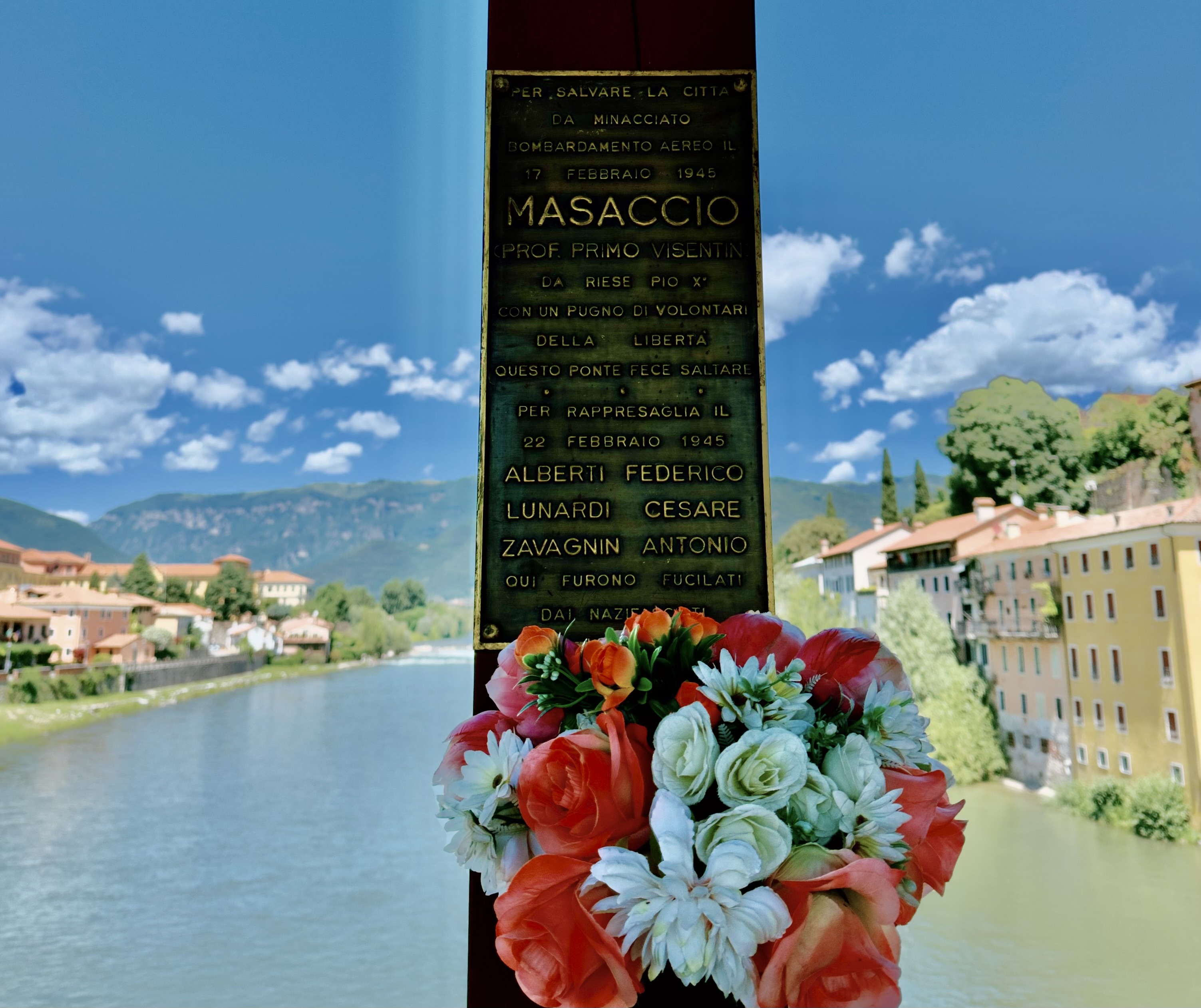 In English: To save the city / from a threatened aerial bombardment, on February 17, 1945 / Masaccio (Prof. Primo Visentin), from Riese Pio X, with a handful of freedom volunteers, blew up this bridge. / In reprisal, on February 22, 1945 / Alberti Federico, Lunardi Cesare, Zavagnin Antonio / were shot here by the Nazi-Fascists. A bronze memorial plaque on a bridge pillar in Bassano del Grappa, decorated with a bouquet of flowers, with the Brenta river and town visible behind it. The plaque commemorates partisan "Masaccio" (Prof. Primo Visentin), who destroyed this bridge on February 17, 1945, to save the city from an air bombardment, and honors three men—Alberti Federico, Lunardi Cesare, and Zavagnin Antonio—who were executed by Nazi-Fascist forces in reprisal on February 22, 1945.