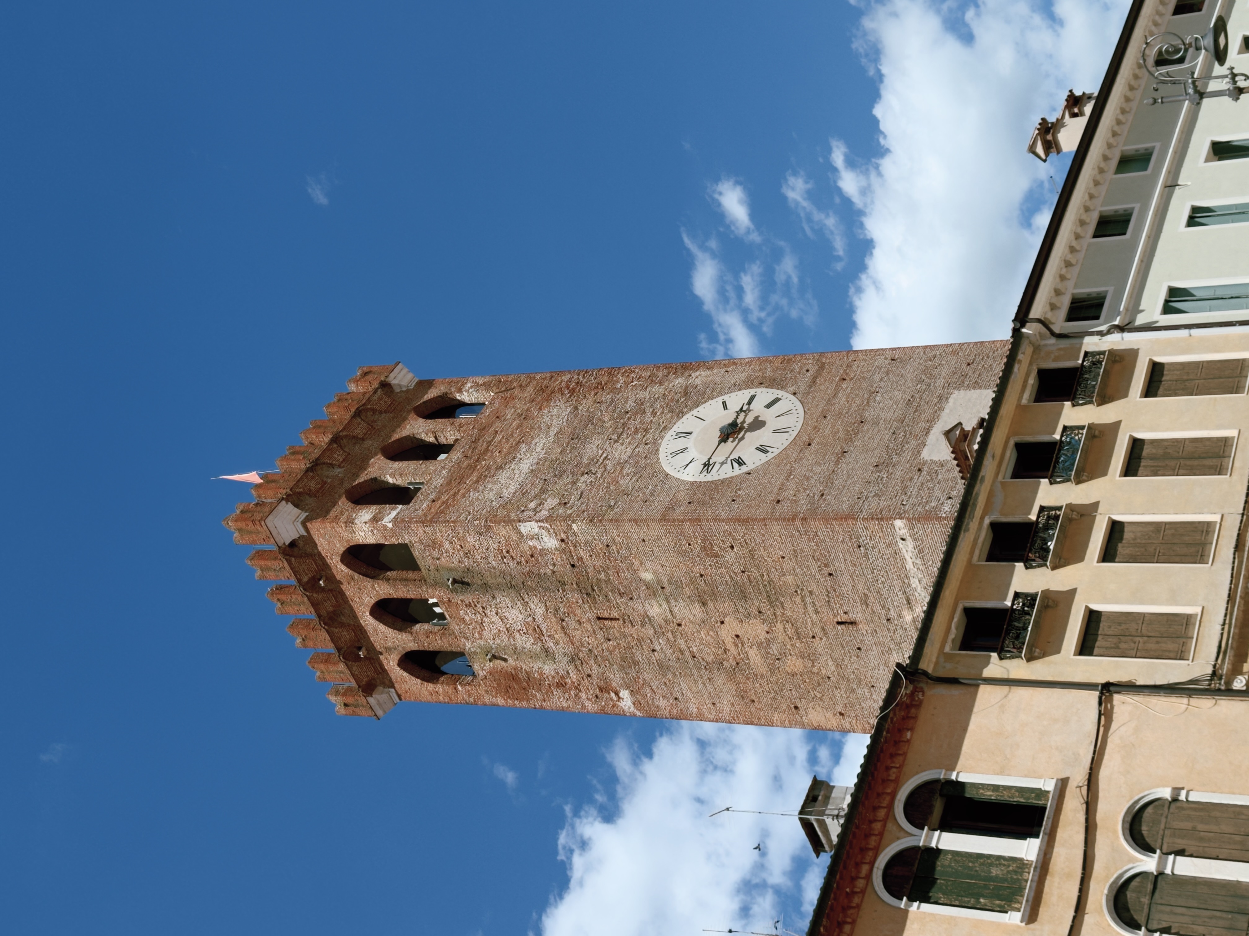 Clock tower in Piazza Garibaldi Looking up at the brick clock tower of Bassano del Grappa's castle, with crenellated battlements and a Roman numeral clock face against a blue sky.