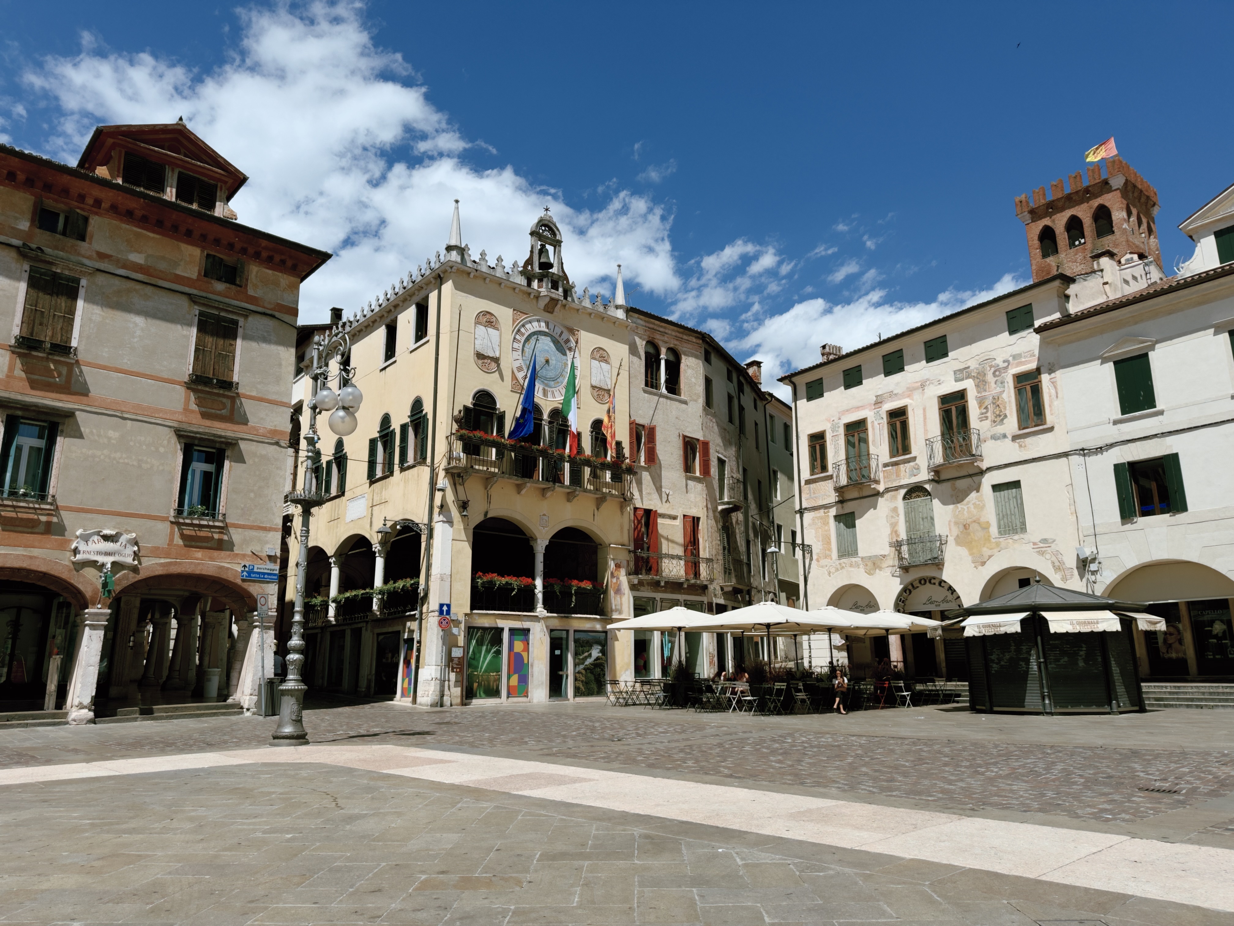 Town hall in Piazza Libertà The town hall of Bassano del Grappa, decorated with EU, Italian, and Veneto flags below its astronomical clock, surrounded by historic frescoed buildings and a brick clock tower in the background.