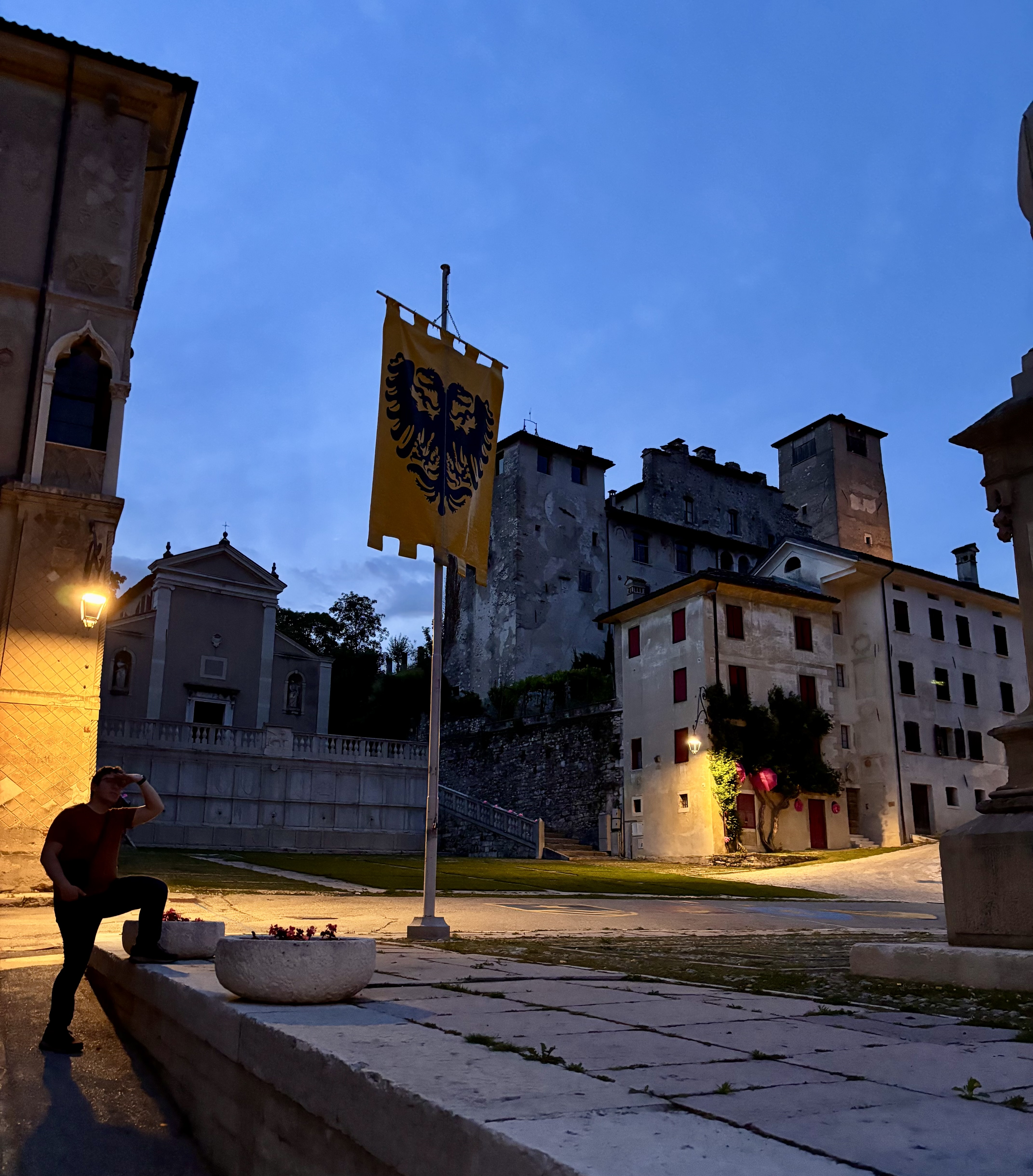 The flag of Port'Oria, last year's Palio di Feltre winner, hangs tall over the town square. A piazza in Feltre at dusk, with a yellow banner bearing a double-headed eagle crest, a church, and the old castle walls and towers in the background.