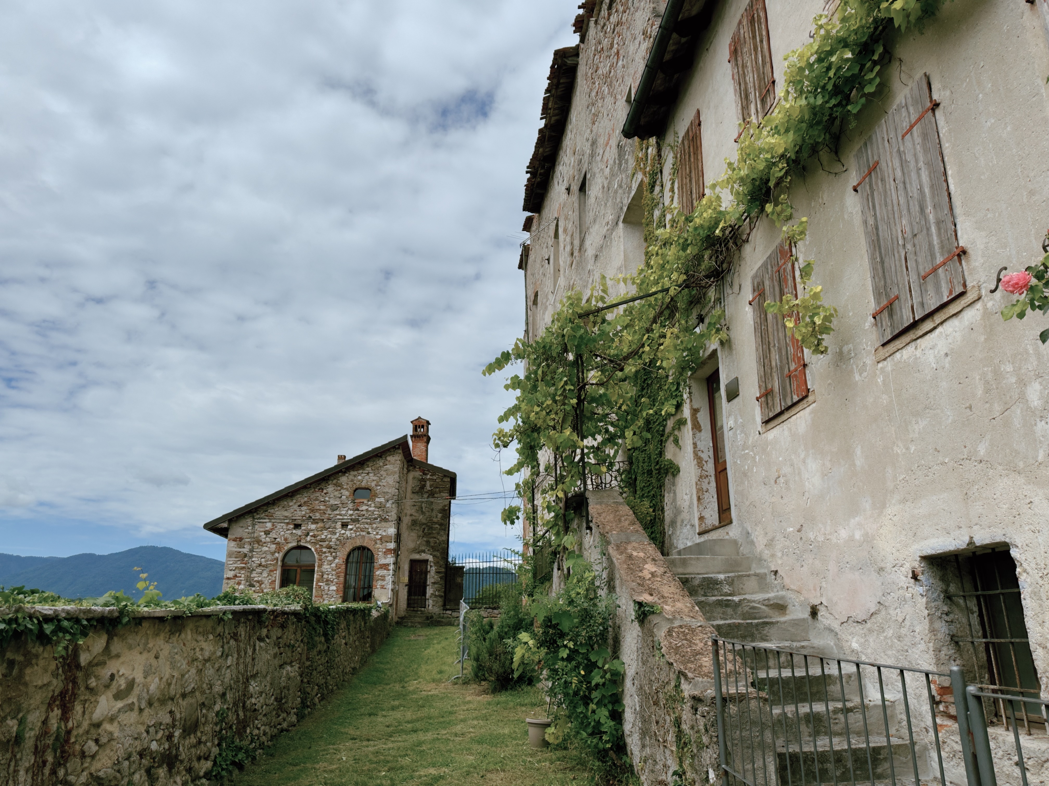 An old stone house in Feltre's hilltop borgo, covered in climbing vines, with stone steps, a grass path, and mountains visible in the distance.