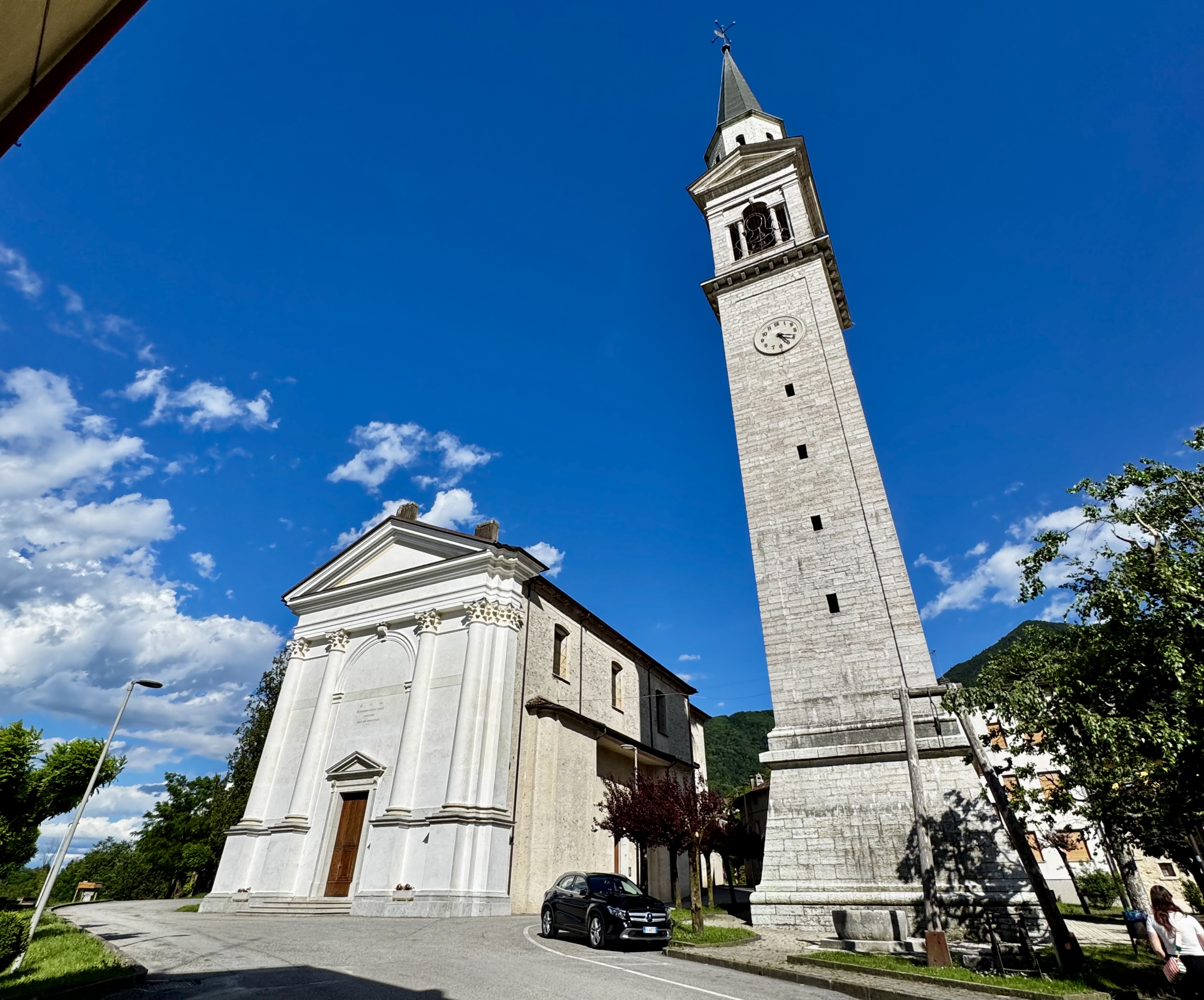 The white church and tall bell tower of Seren del Grappa against a clear blue sky, with green hills in the background.