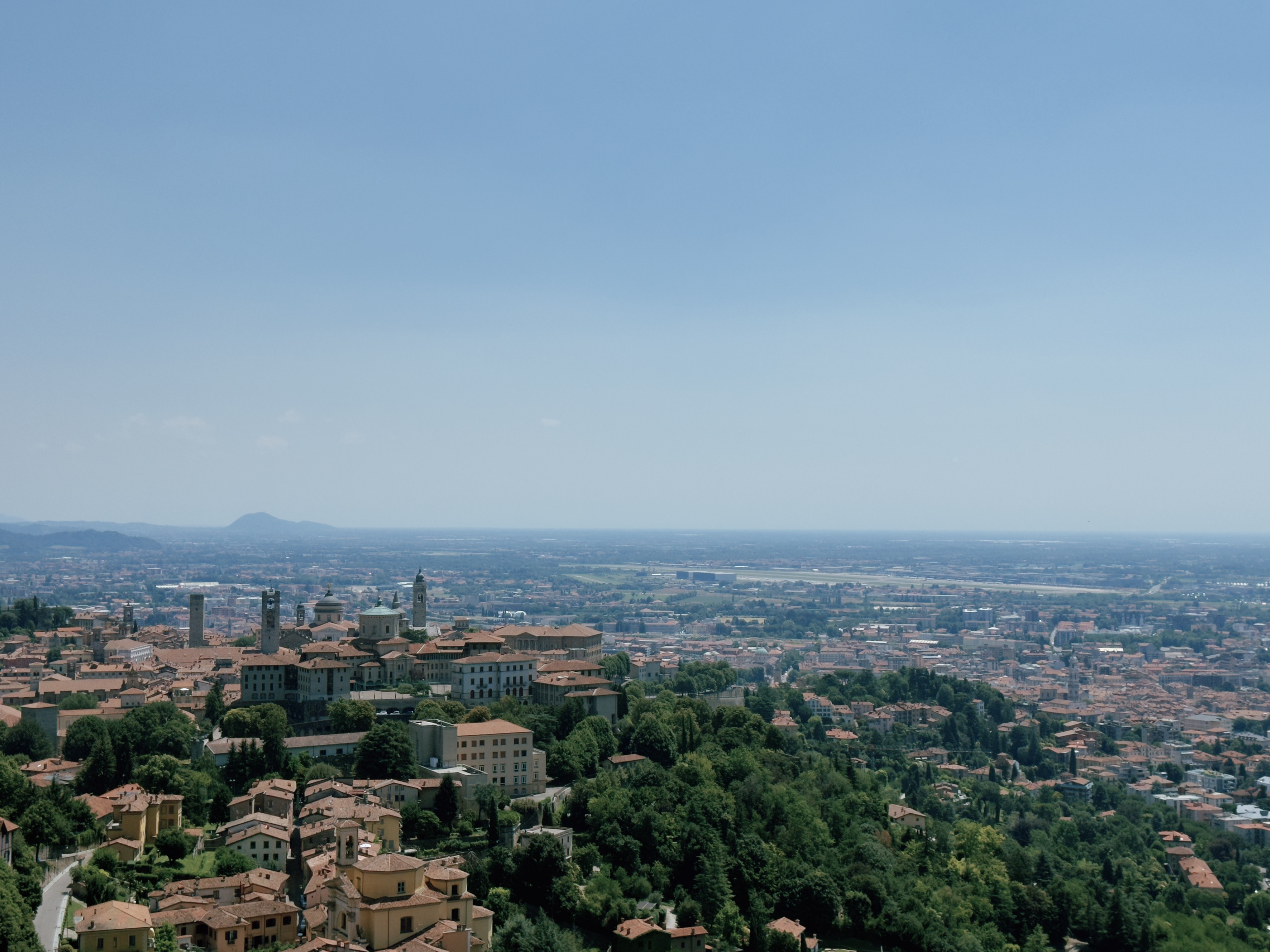 Aerial view of Bergamo from Città Alta, with the historic upper town's towers and cathedral domes in the foreground and the flat Po Valley stretching to the horizon.