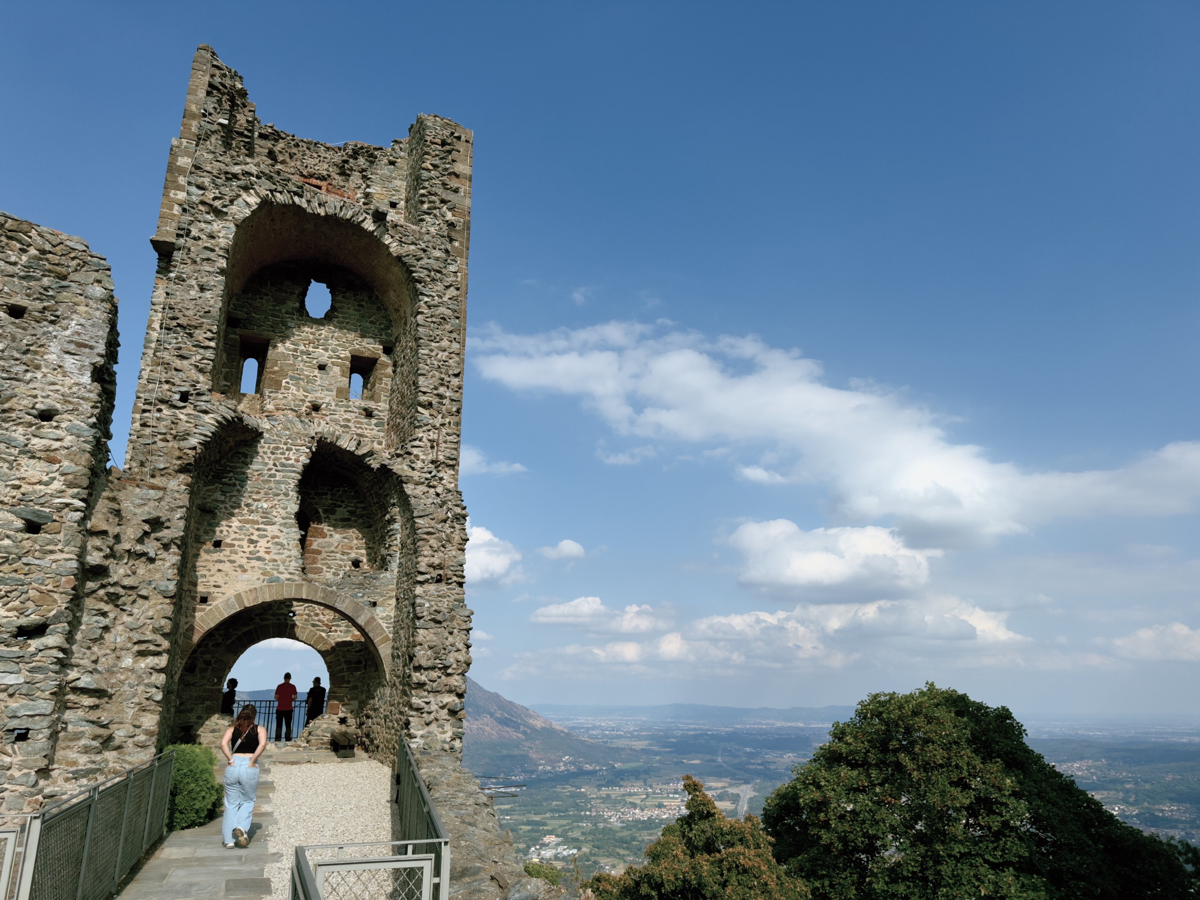 Visitors walking through the ruined stone tower of the Sacra di San Michele abbey, known as the Torre della Bell'Alda, with sweeping views of the Piedmont valley beyond.