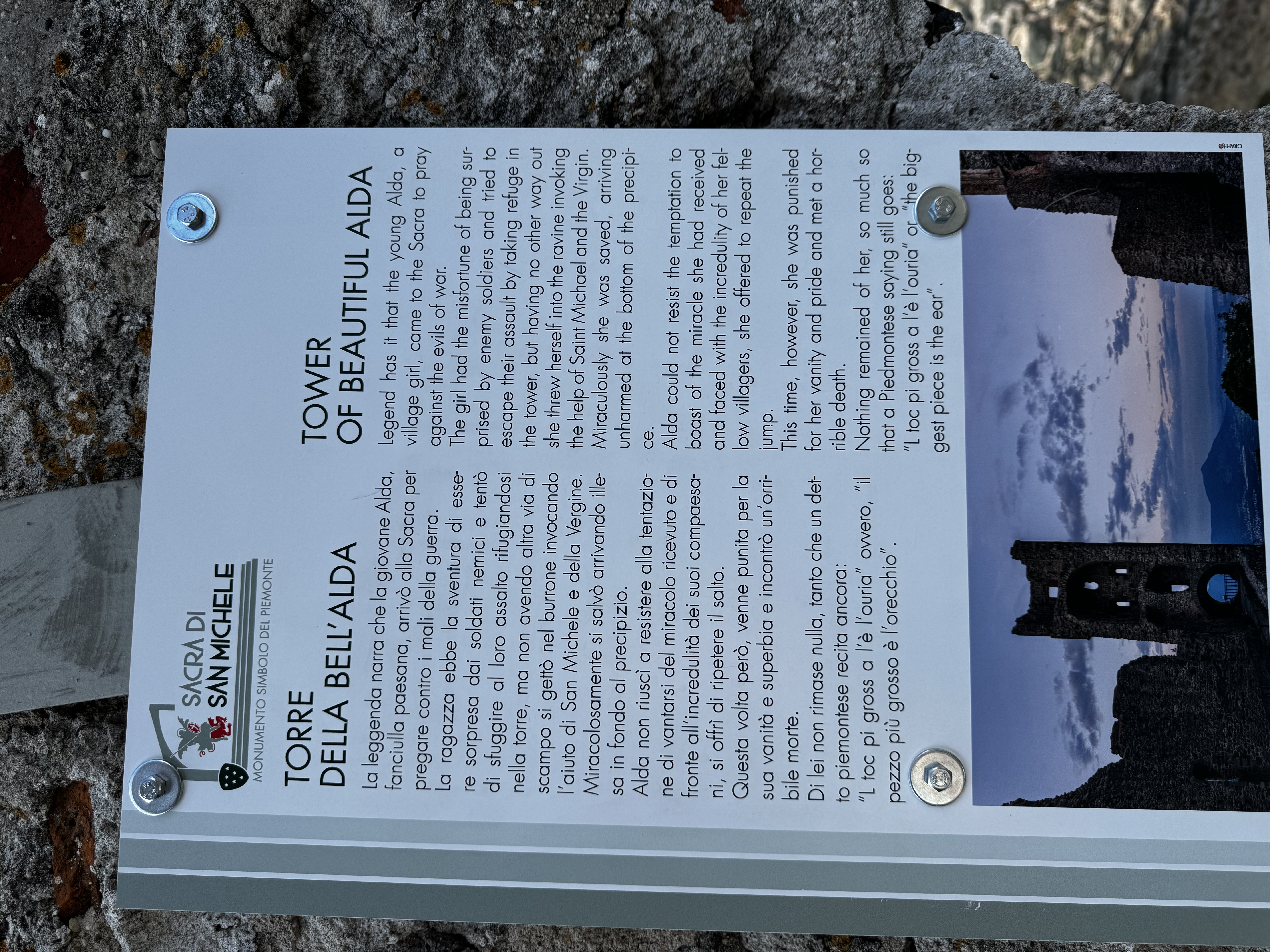 An information plaque at Sacra di San Michele telling, in Italian and English, the legend of the Torre della Bell'Alda: a village girl named Alda, fleeing enemy soldiers, jumped from the tower and survived unharmed after invoking Saint Michael and the Virgin, but died when she vainly tried to repeat the jump to prove the miracle to disbelieving villagers. Illustrated with a photo of the tower at dusk.