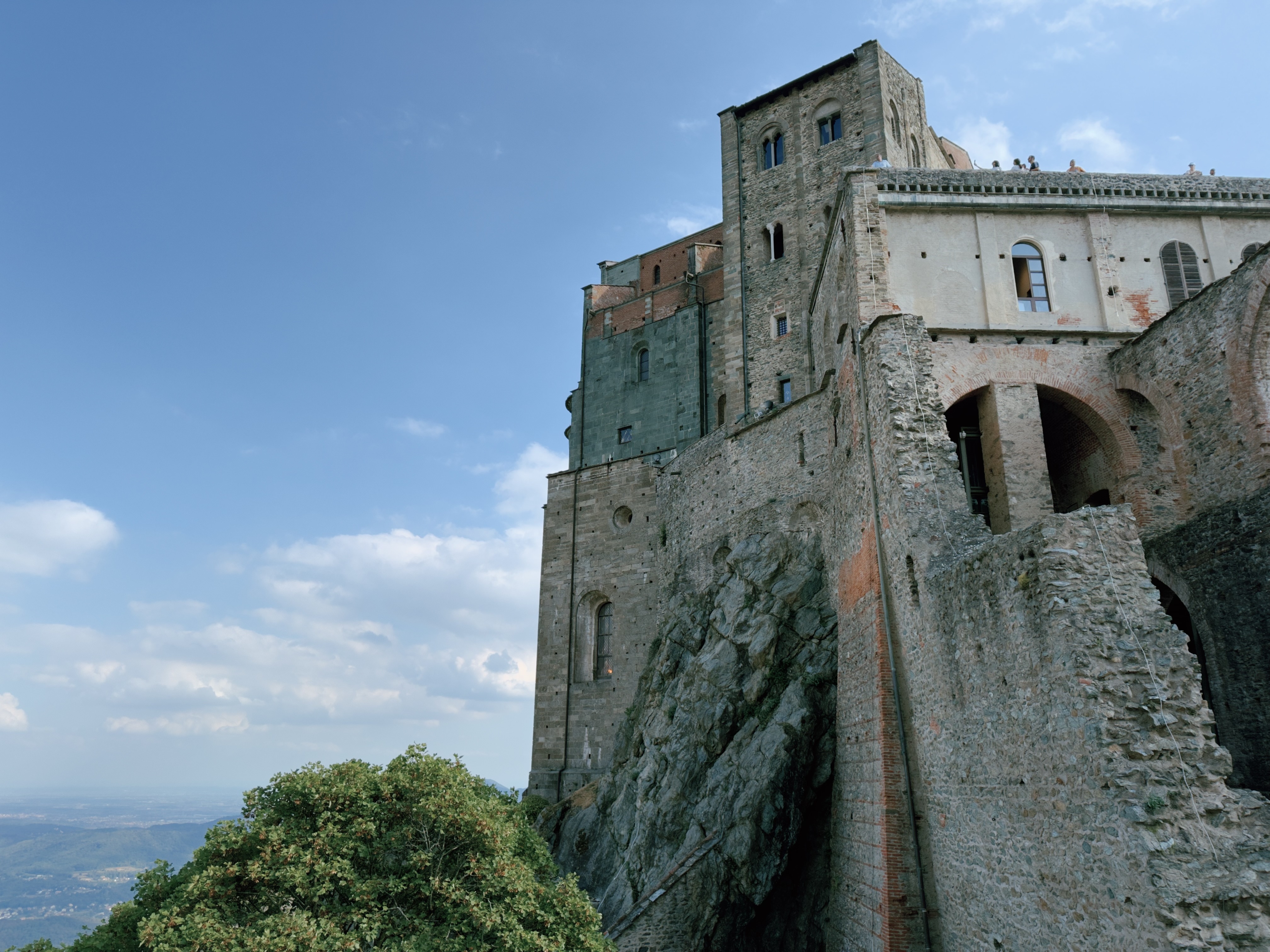 The medieval abbey of Sacra di San Michele in Piedmont, its stone walls and tower built directly onto a rocky mountain outcrop, with visitors viewing from the terrace above.