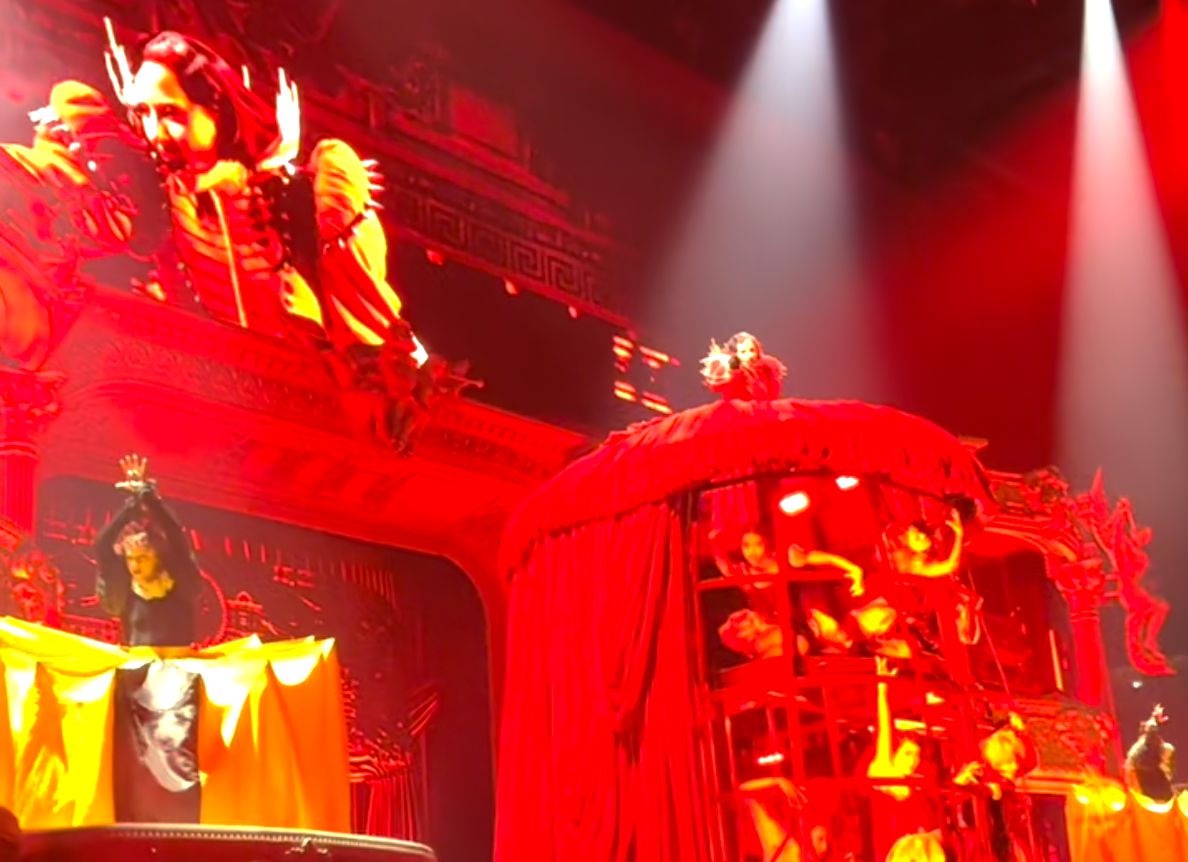 Lady Gaga on stage. She stands on top of a metal cage covered in a giant red dress, and points at the camera. There's a lot of people inside that cage