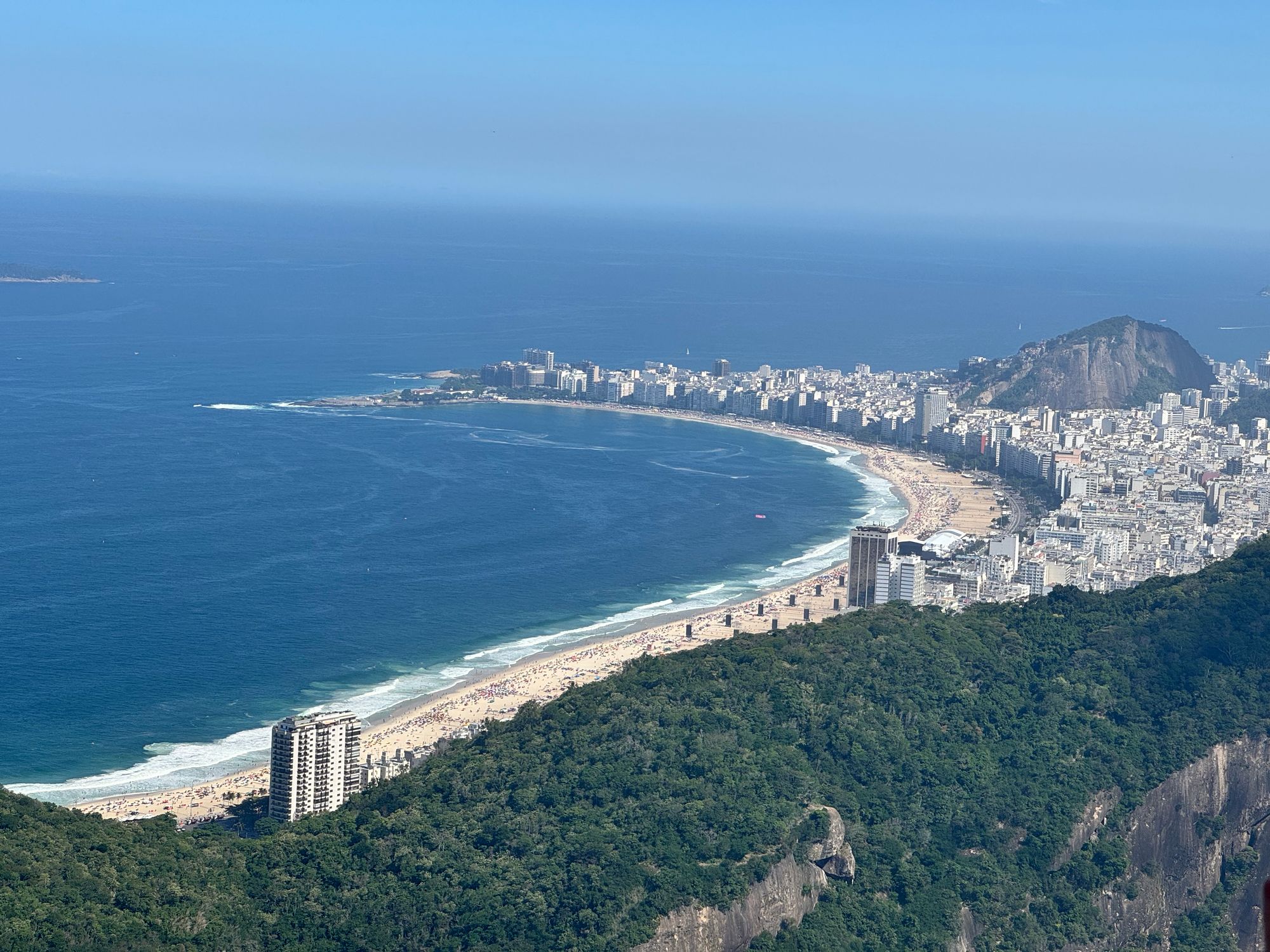 Picture of Copacabana beach seen from above. Weather is sunny, sky and see are blue. The beach is curved and surrounded by tall buildings, which are themselves surrounded by native atlantic forests.