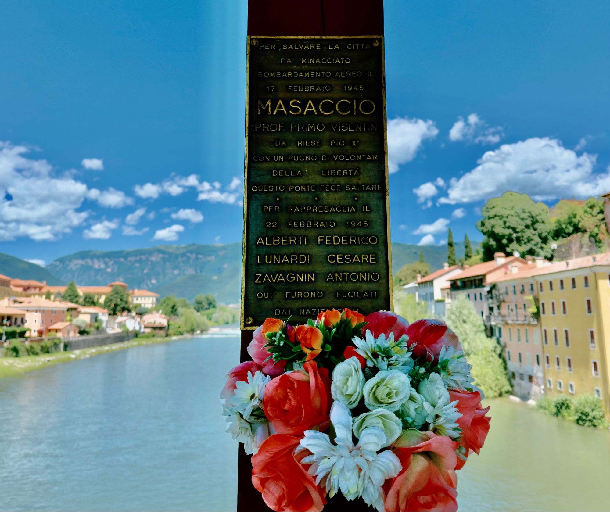 A bronze memorial plaque on a bridge pillar in Bassano del Grappa, decorated with a bouquet of flowers, with the Brenta river and town visible behind it. The plaque commemorates partisan "Masaccio" (Prof. Primo Visentin), who destroyed this bridge on February 17, 1945, to save the city from an air bombardment, and honors three men—Alberti Federico, Lunardi Cesare, and Zavagnin Antonio—who were executed by Nazi-Fascist forces in reprisal on February 22, 1945.
