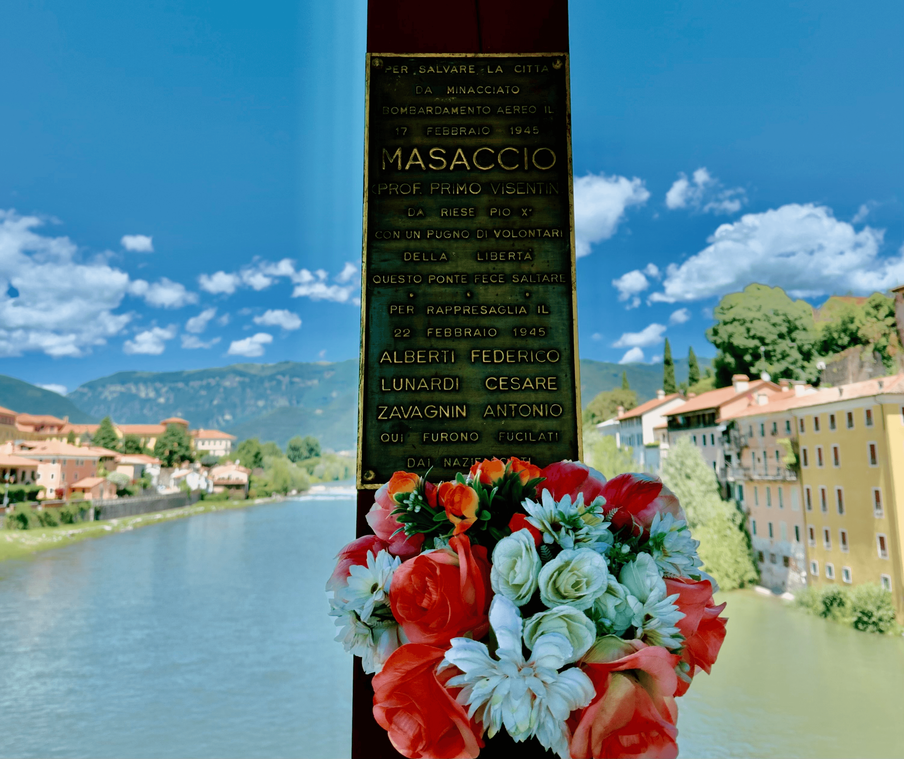 A bronze memorial plaque on a bridge pillar in Bassano del Grappa, decorated with a bouquet of flowers, with the Brenta river and town visible behind it. The plaque commemorates partisan "Masaccio" (Prof. Primo Visentin), who destroyed this bridge on February 17, 1945, to save the city from an air bombardment, and honors three men—Alberti Federico, Lunardi Cesare, and Zavagnin Antonio—who were executed by Nazi-Fascist forces in reprisal on February 22, 1945.