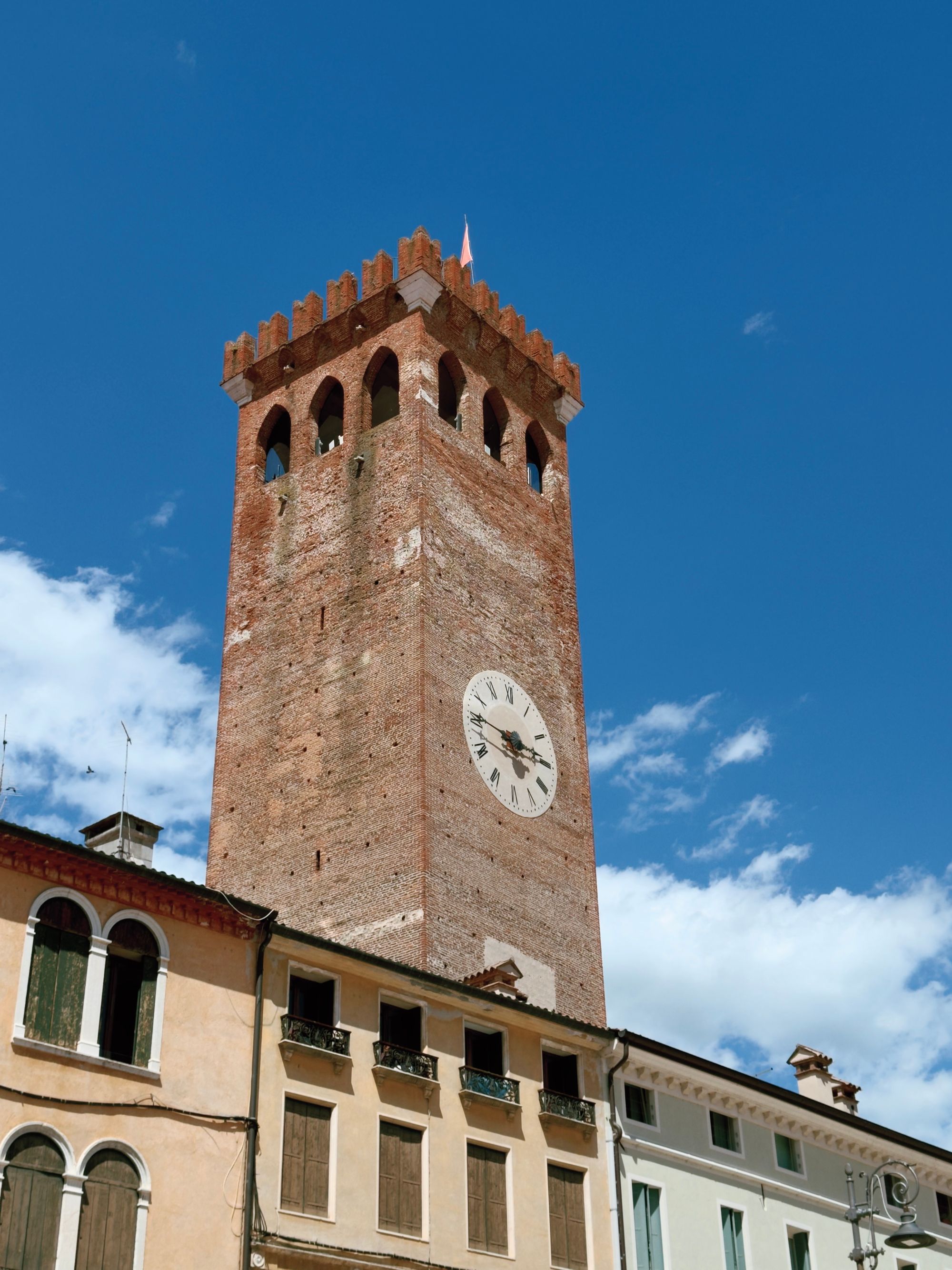 Looking up at the brick clock tower of Bassano del Grappa's castle, with crenellated battlements and a Roman numeral clock face against a blue sky.