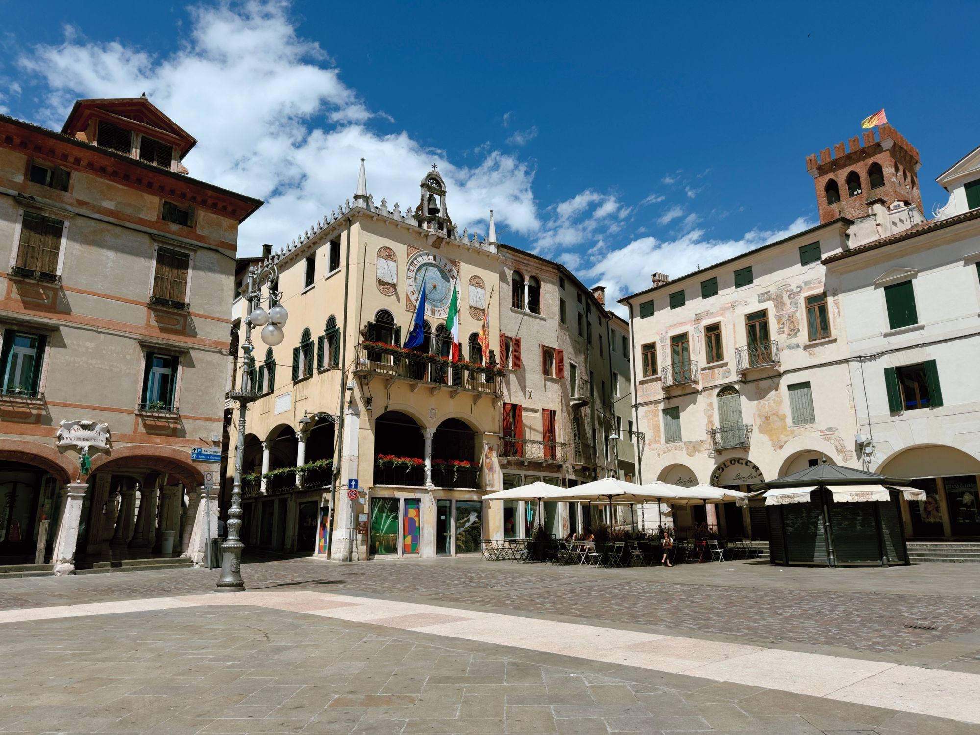 The town hall of Bassano del Grappa, decorated with EU, Italian, and Veneto flags below its astronomical clock, surrounded by historic frescoed buildings and a brick clock tower in the background.