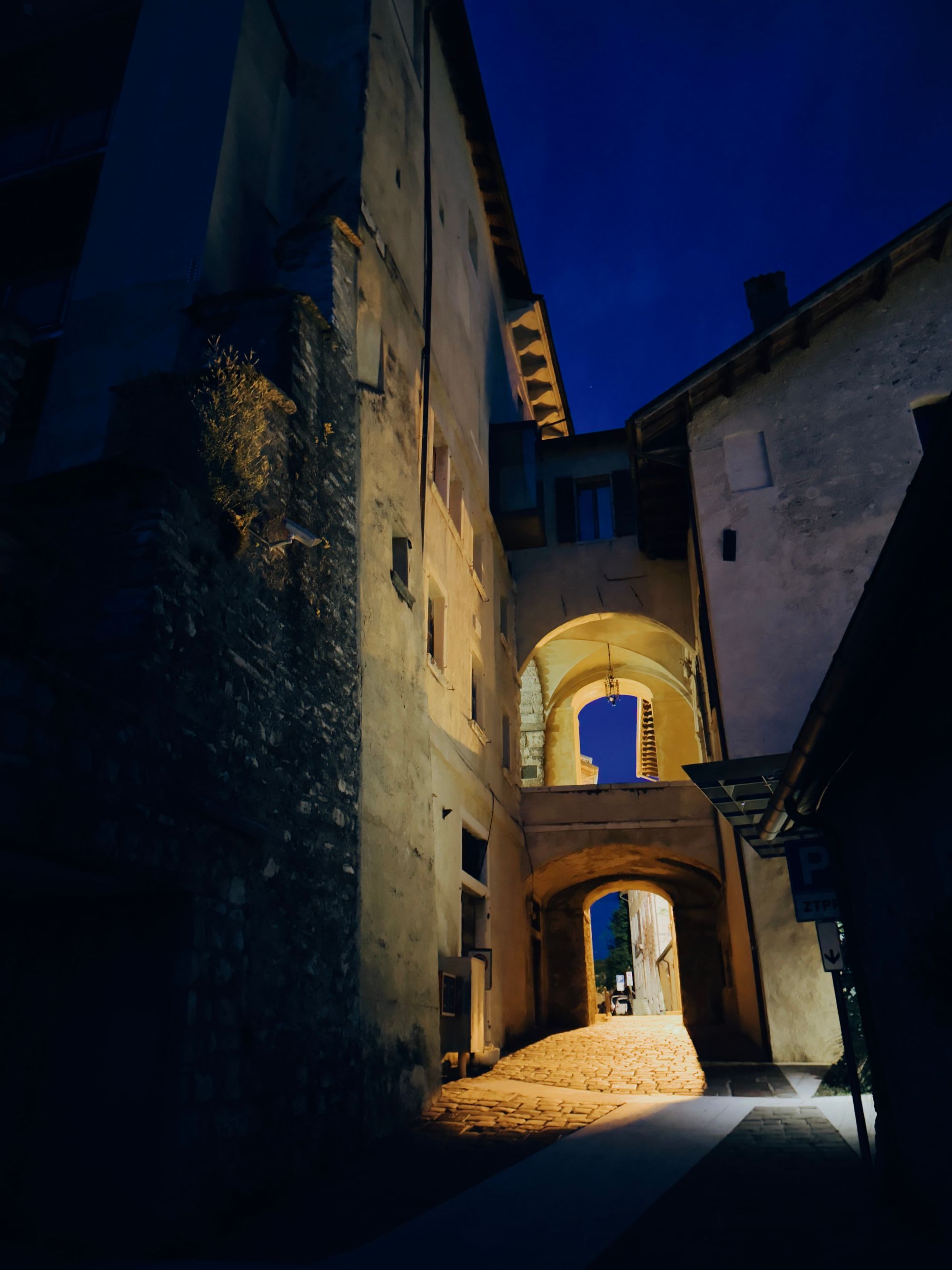 A narrow cobblestone alley in Feltre at night, passing under stone archways between old buildings, lit by warm light against a deep blue sky.