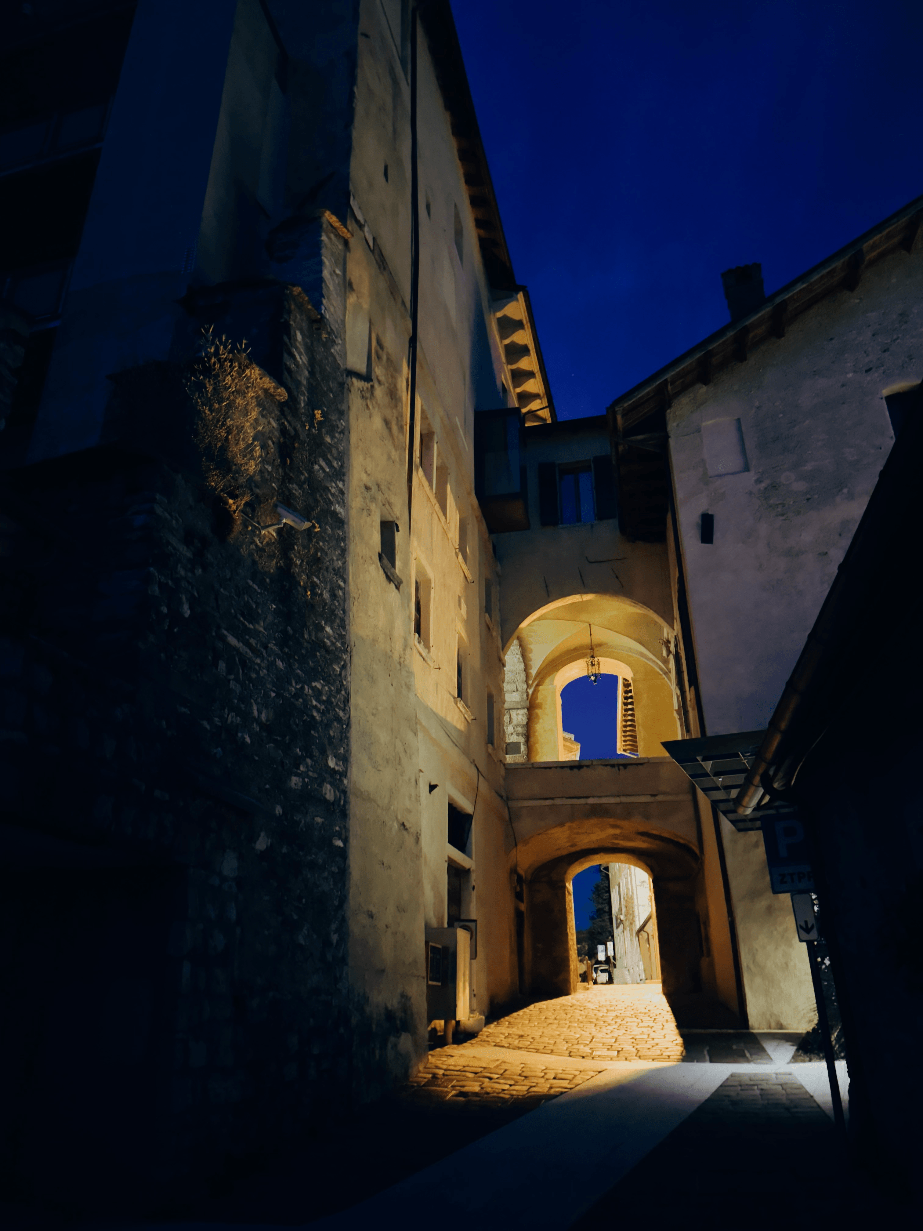 A narrow cobblestone alley in Feltre at night, passing under stone archways between old buildings, lit by warm light against a deep blue sky.
