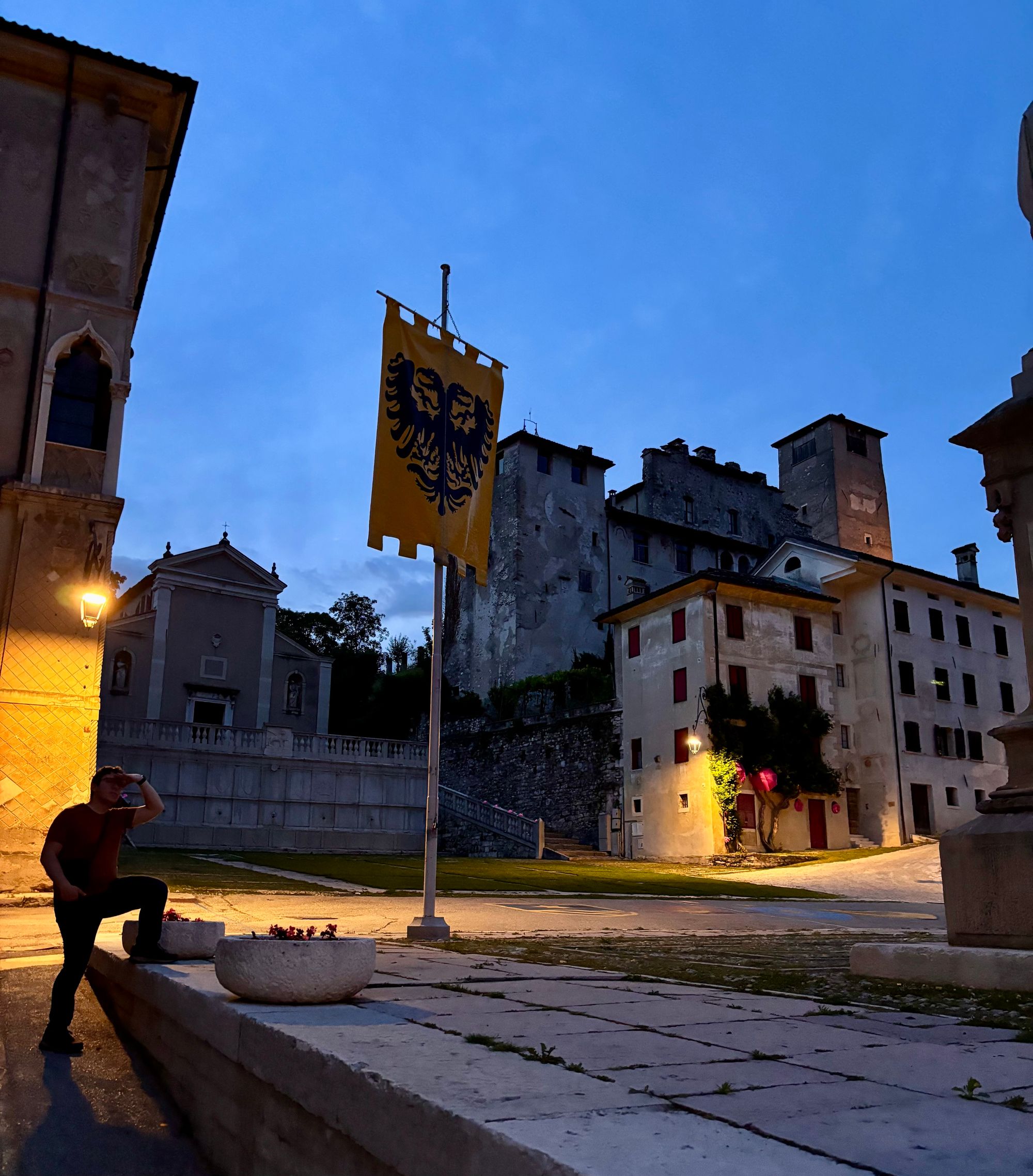 A piazza in Feltre at dusk, with a yellow banner bearing a double-headed eagle crest, a church, and the old castle walls and towers in the background.