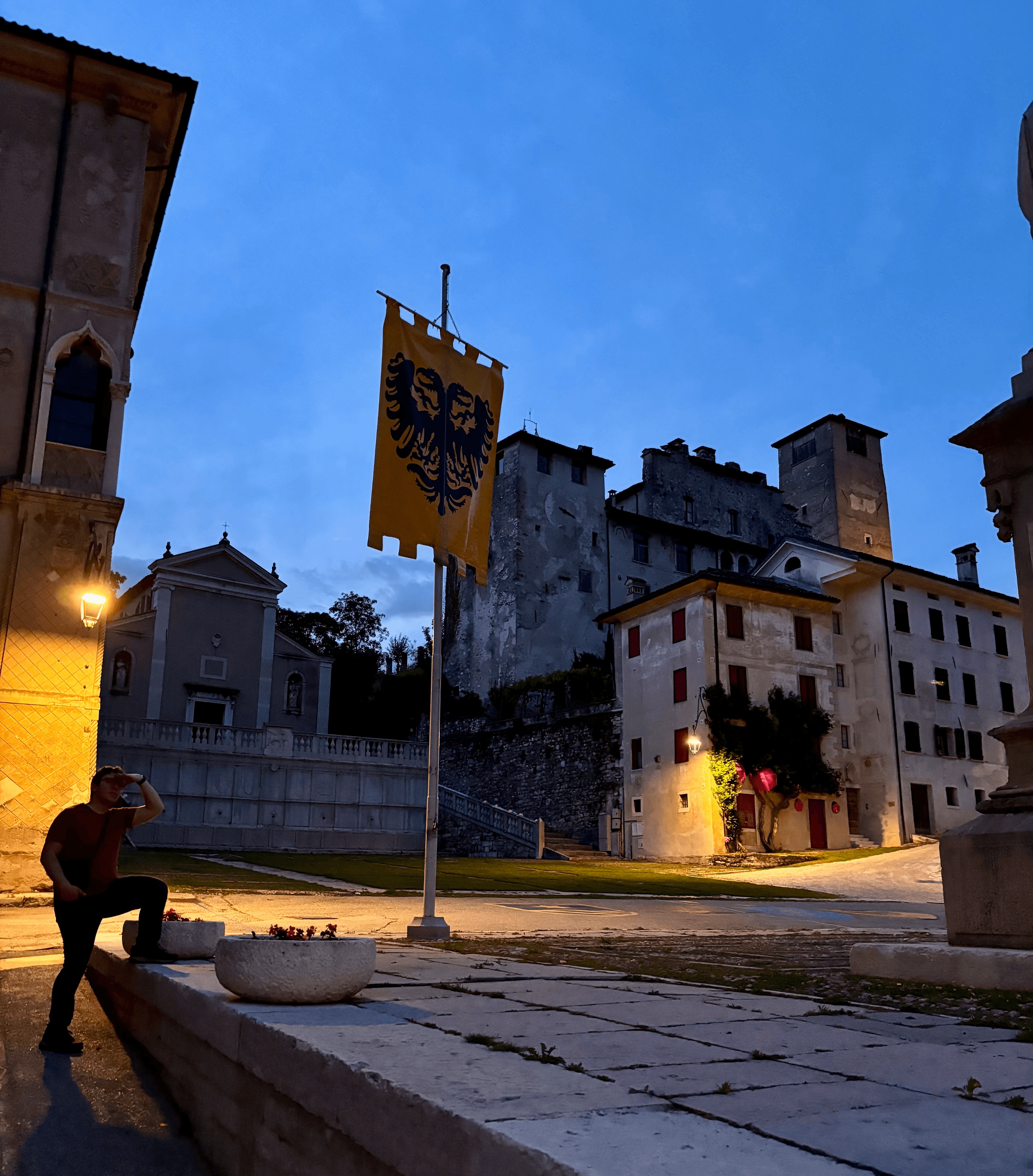 A piazza in Feltre at dusk, with a yellow banner bearing a double-headed eagle crest, a church, and the old castle walls and towers in the background.