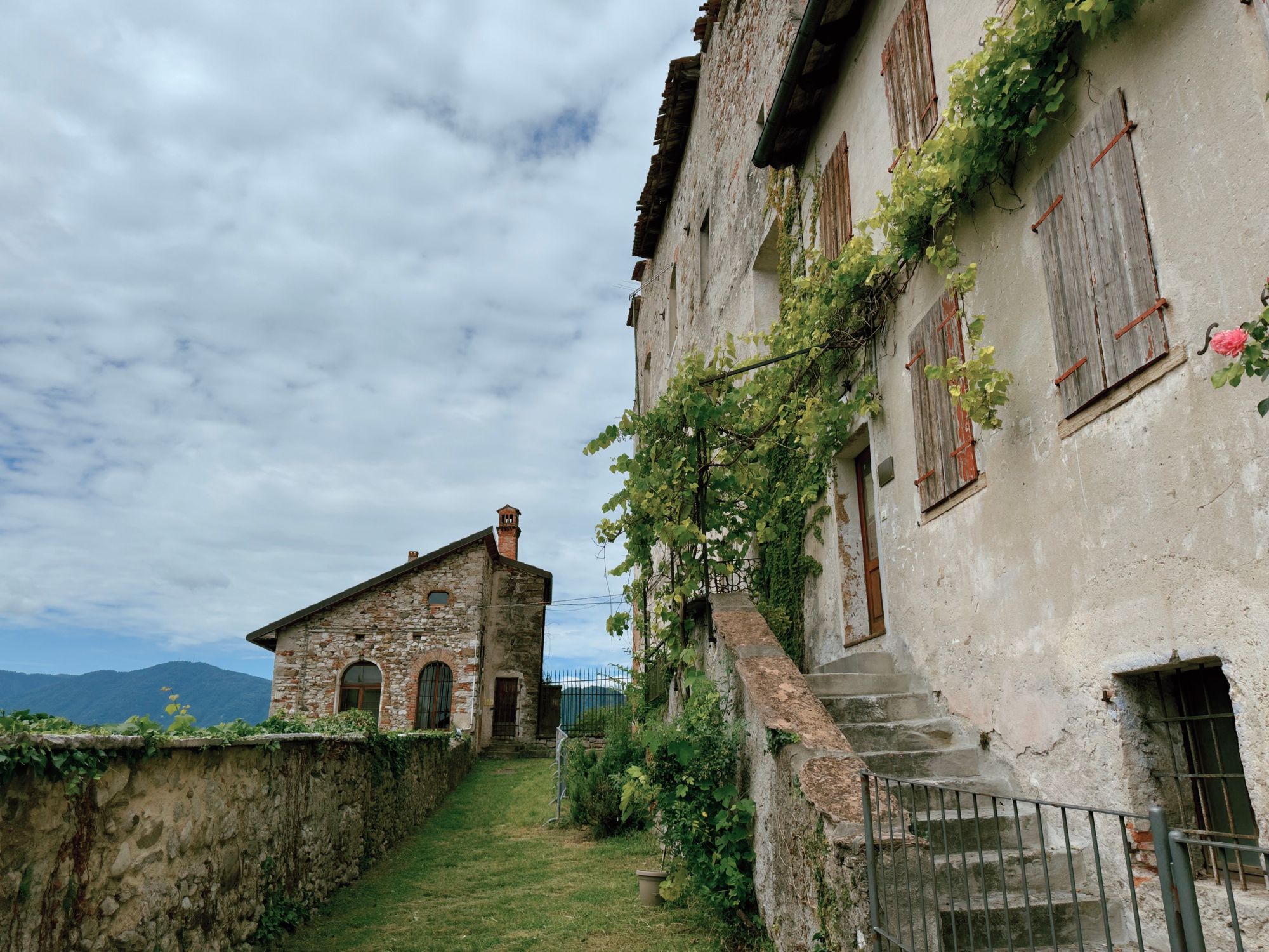 An old stone house in Feltre's hilltop borgo, covered in climbing vines, with stone steps, a grass path, and mountains visible in the distance.