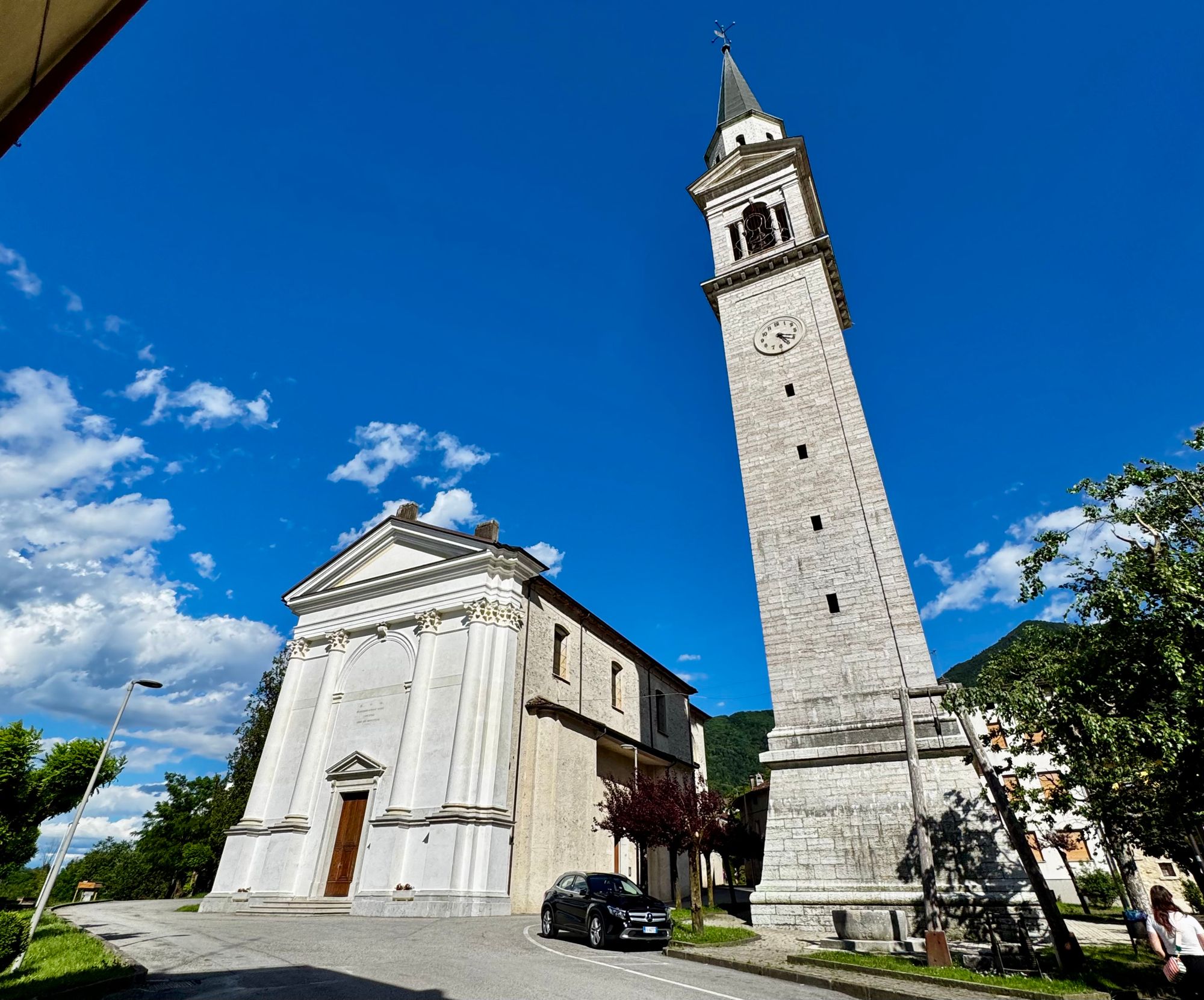 The white church and tall bell tower of Seren del Grappa against a clear blue sky, with green hills in the background.