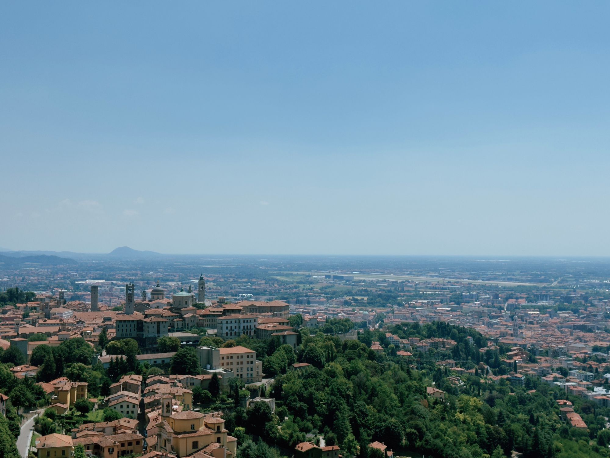 Aerial view of Bergamo from Città Alta, with the historic upper town's towers and cathedral domes in the foreground and the flat Po Valley stretching to the horizon.