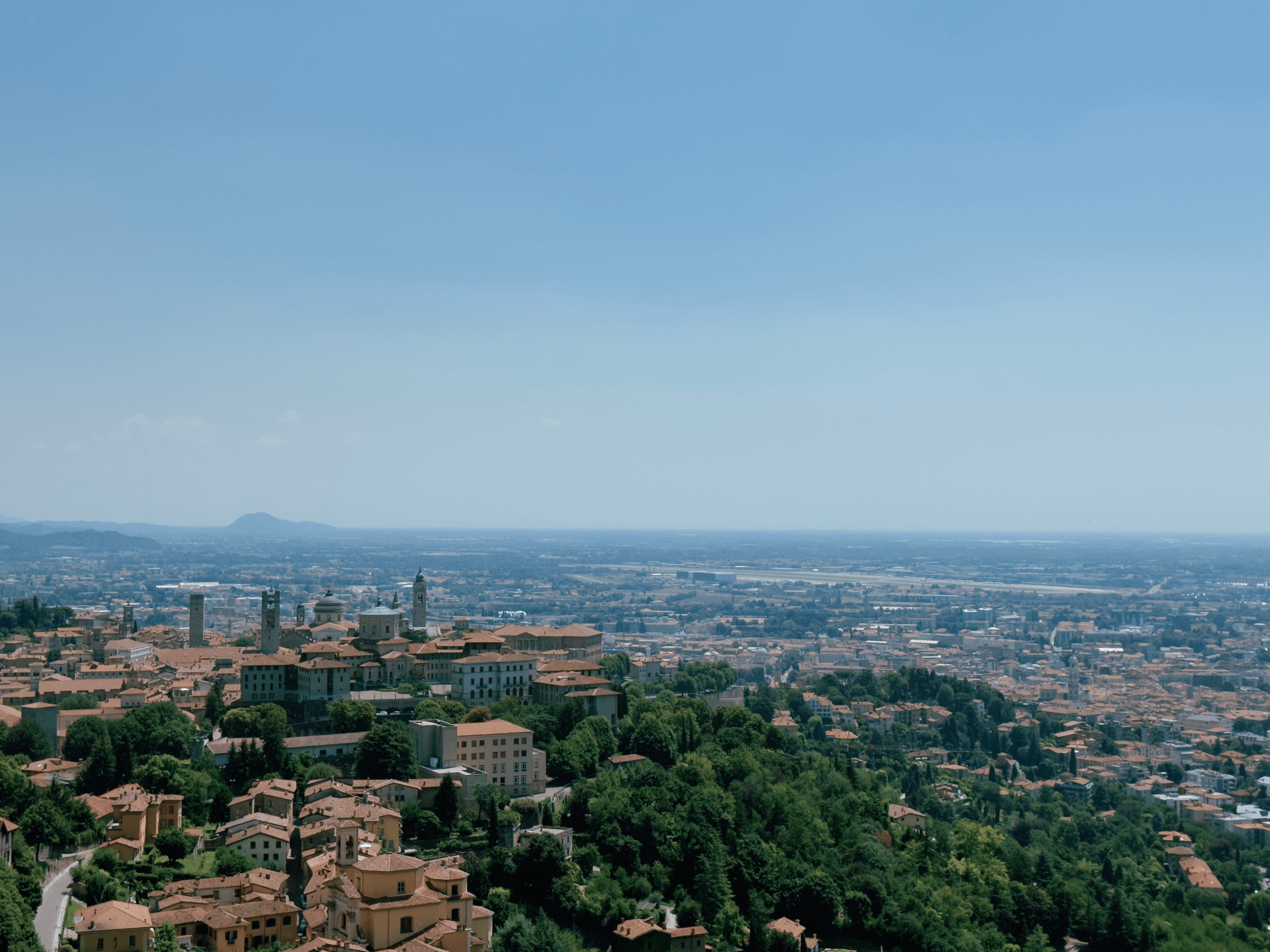 Aerial view of Bergamo from Città Alta, with the historic upper town's towers and cathedral domes in the foreground and the flat Po Valley stretching to the horizon.