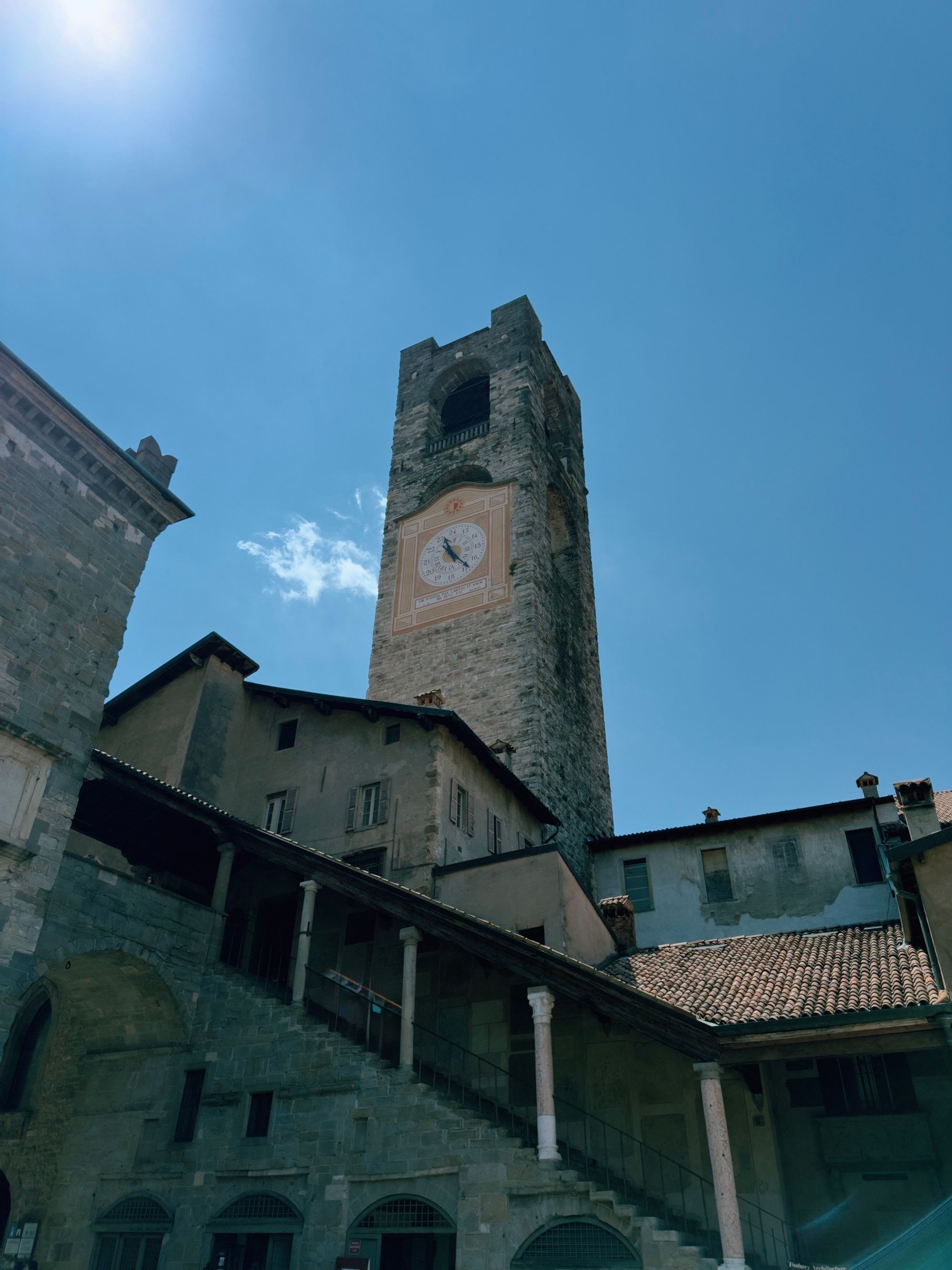 Looking up at the Campanone, Bergamo's medieval stone bell tower in Piazza Vecchia, with its decorative clock face and open bell chamber against a bright blue sky.