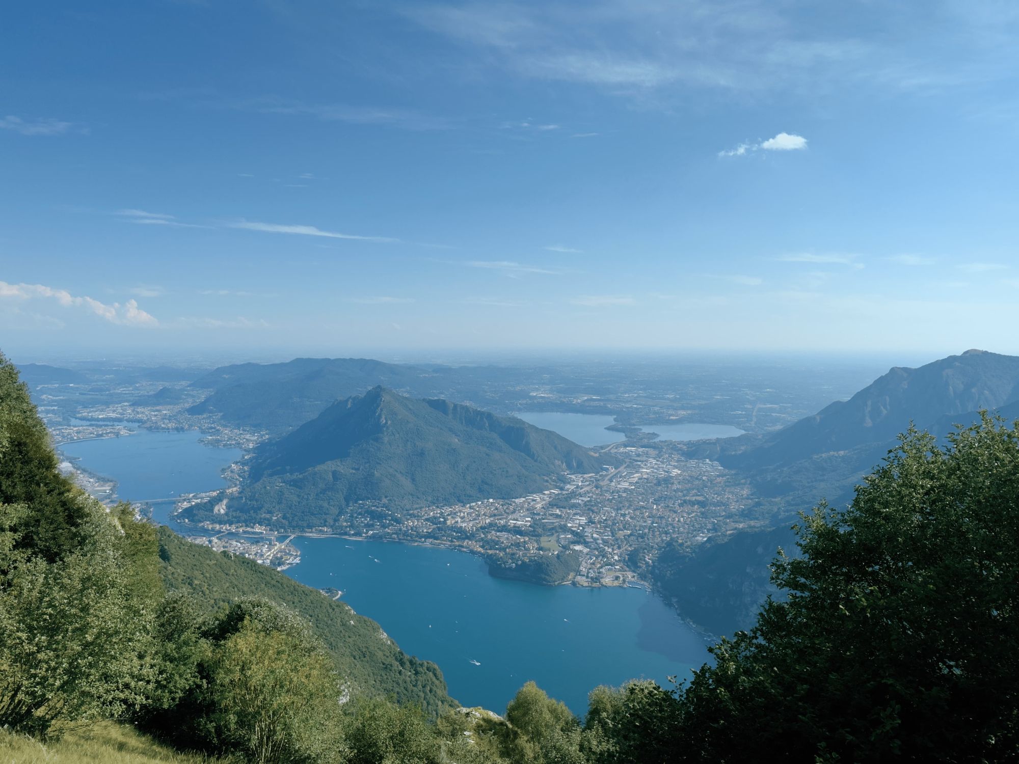 Aerial view from Monte Coltignone looking down over Lake Como and the city of Valmadrera, with the branches of the lake and surrounding mountains stretching into the hazy distance.