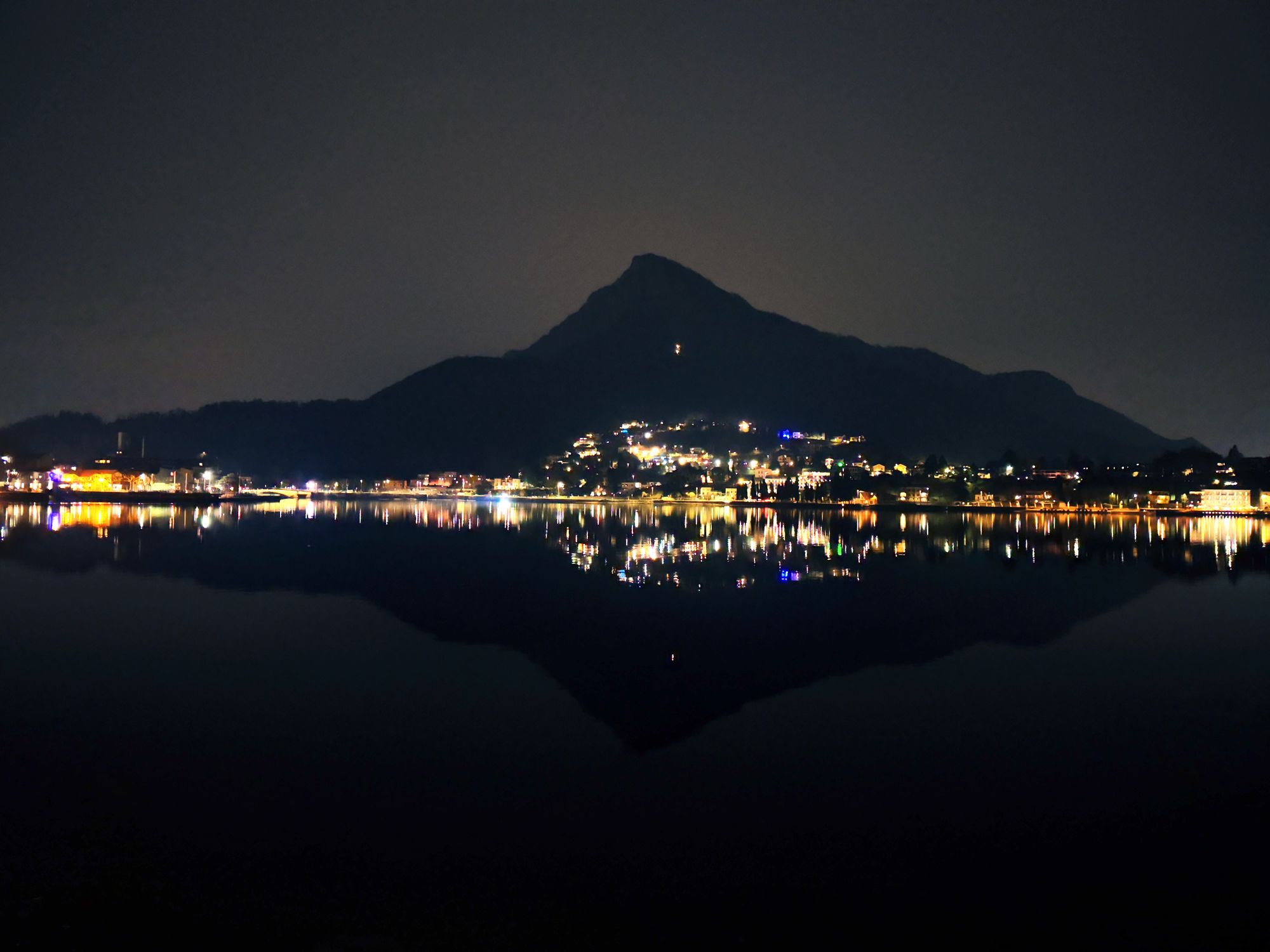 The lights of a city at night and the shadow of the mountain behind it are perfectly reflected in the dark waters of the lake.