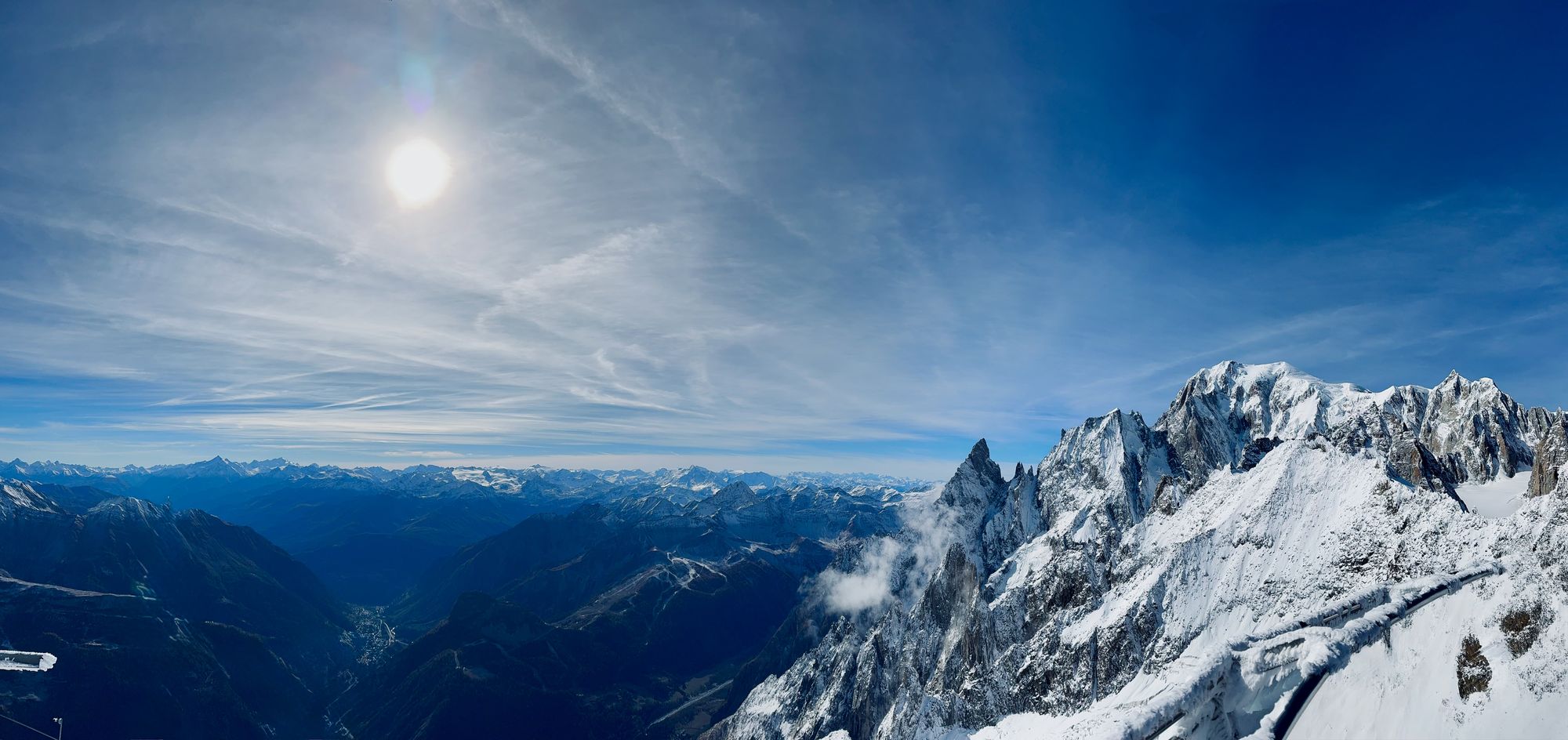 Panoramic view from Punta Helbronner, with jagged granite spires in the foreground, Mont Blanc's snow-covered peaks on the right, and a hazy sun over a sea of distant alpine ranges.
