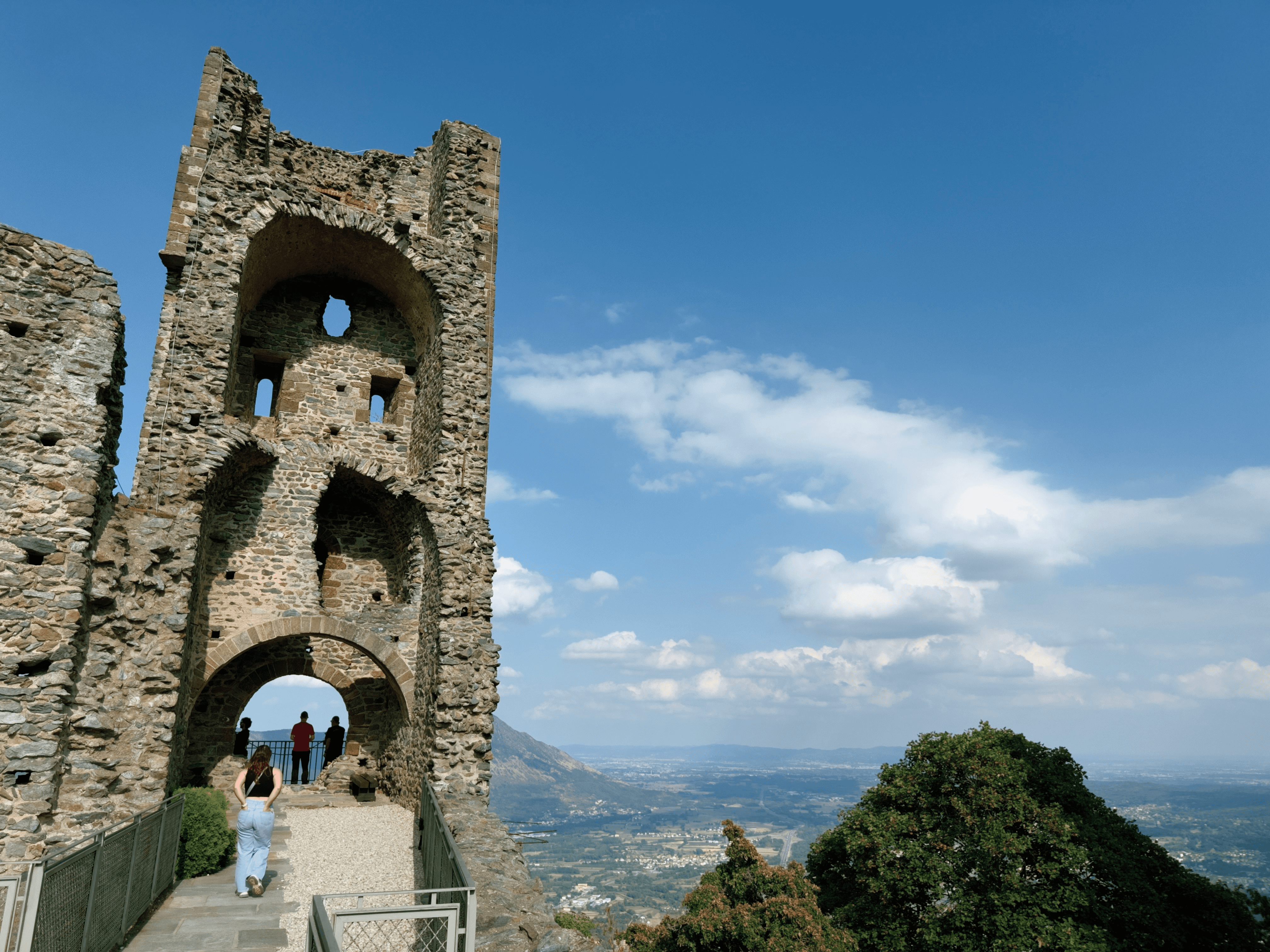 Visitors walking through the ruined stone tower of the Sacra di San Michele abbey, known as the Torre della Bell'Alda, with sweeping views of the Piedmont valley beyond.
