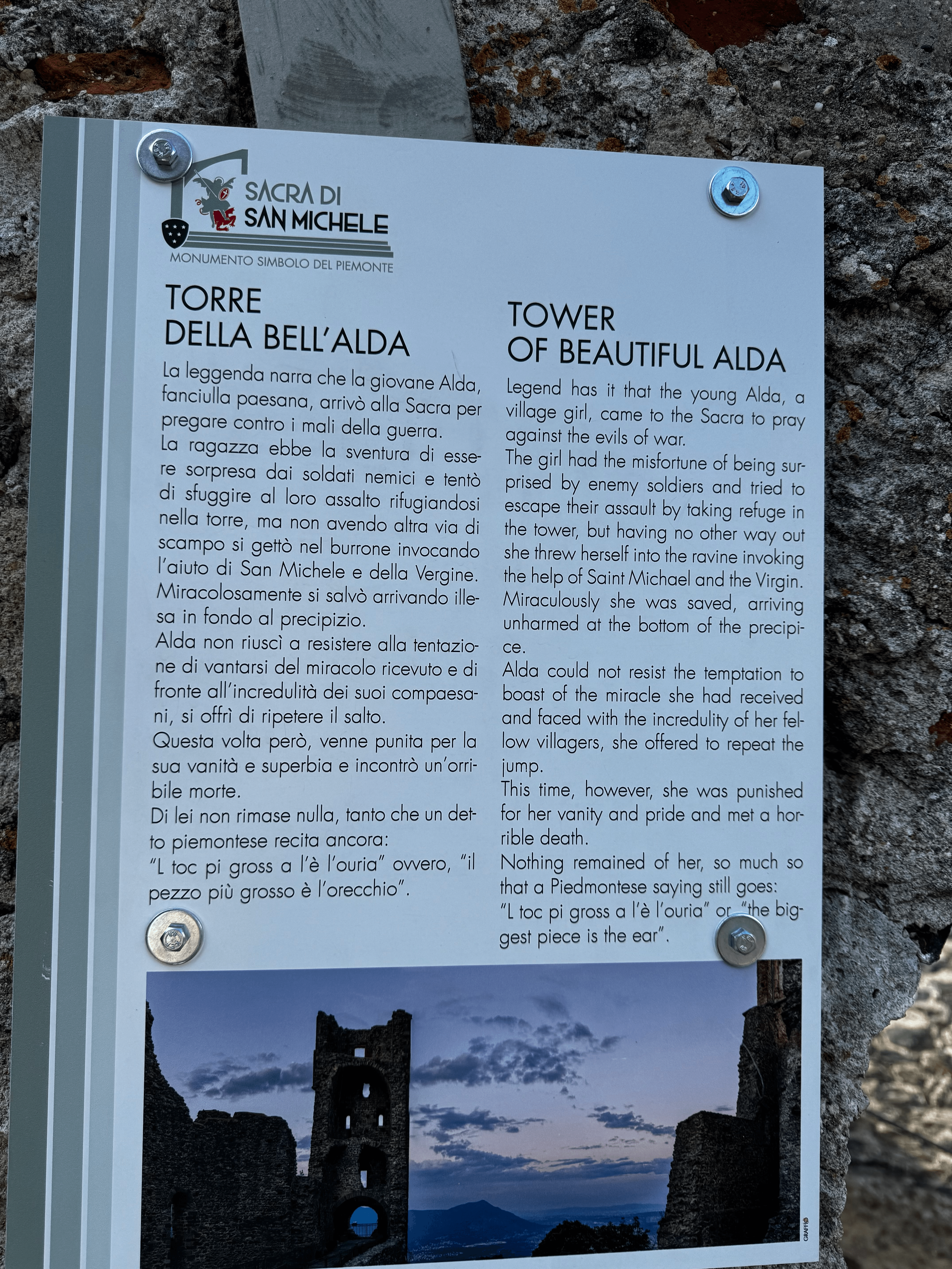 An information plaque at Sacra di San Michele telling, in Italian and English, the legend of the Torre della Bell'Alda: a village girl named Alda, fleeing enemy soldiers, jumped from the tower and survived unharmed after invoking Saint Michael and the Virgin, but died when she vainly tried to repeat the jump to prove the miracle to disbelieving villagers. Illustrated with a photo of the tower at dusk.