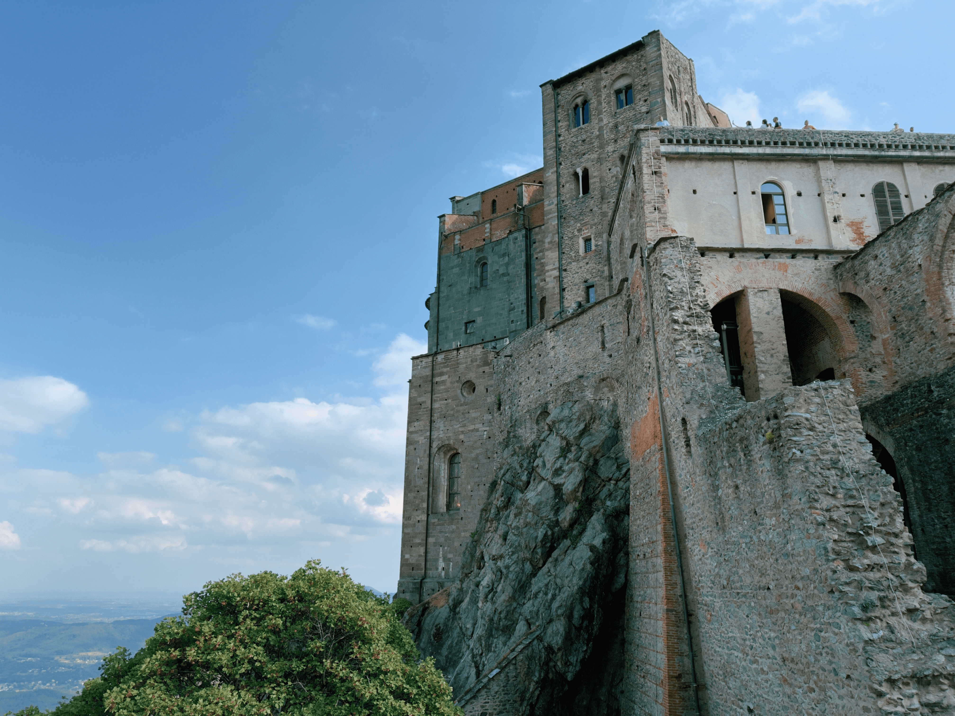 The medieval abbey of Sacra di San Michele in Piedmont, its stone walls and tower built directly onto a rocky mountain outcrop, with visitors viewing from the terrace above.