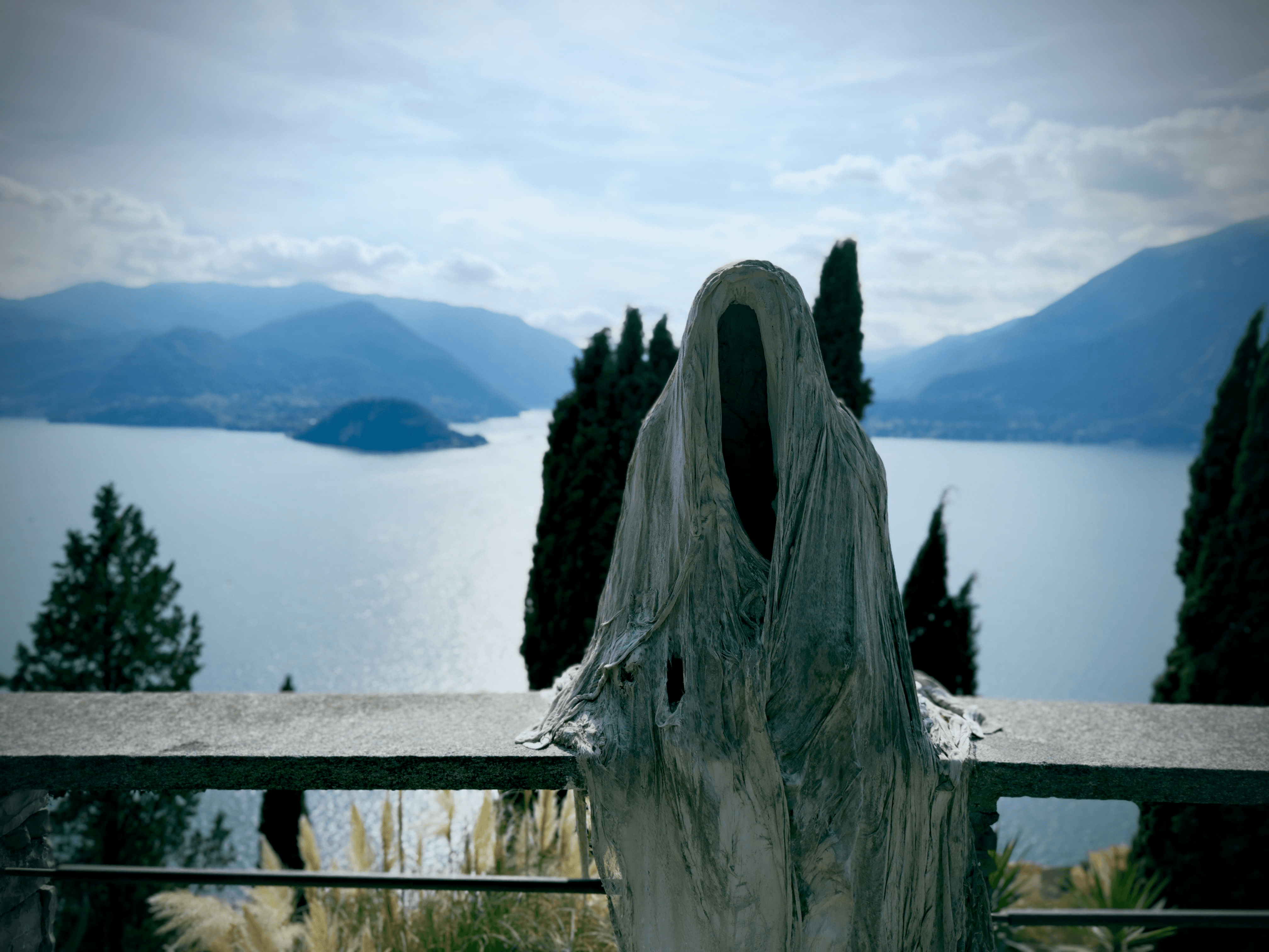 A ghostly hooded figure sculpture standing on a terrace overlooking Lake Como near Vezio, with cypress trees and misty mountains framing the view.